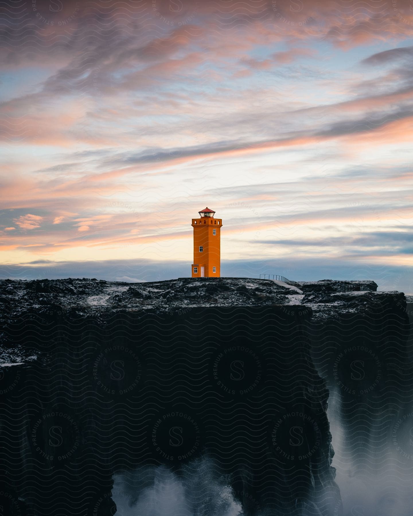 A lighthouse sits at the edge of a coastal cliff under pink clouds at dusk.