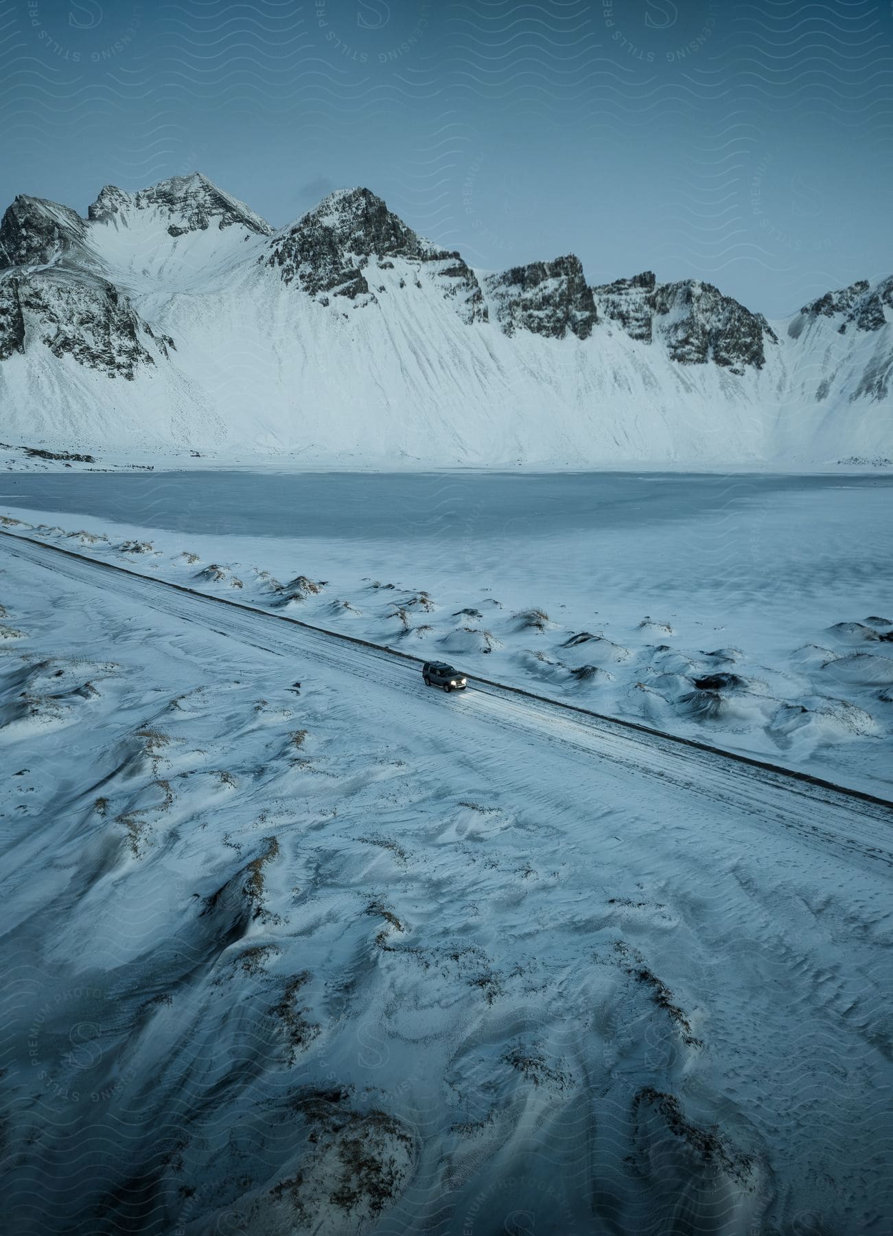 An aerial view of a car driving down a road near some tall mountains covered in snow.