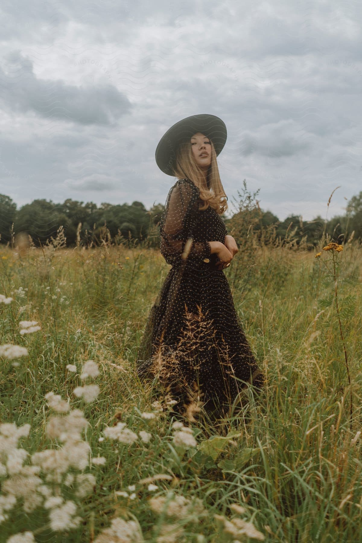 A woman in a black polka dot dress and hat stands on a grassy field with blooming flowers in the foreground on a cloudy day, her shoulder-length blonde hair curling up at the ends.