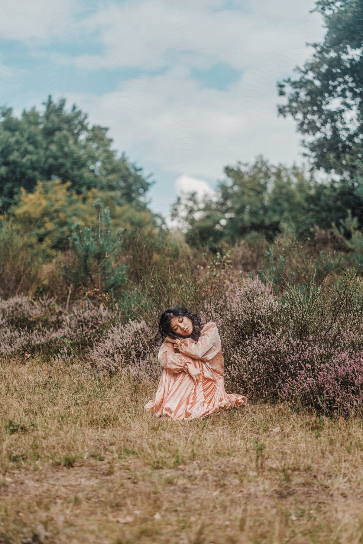 Woman sitting in a lush environment wearing a long pink satin dress.