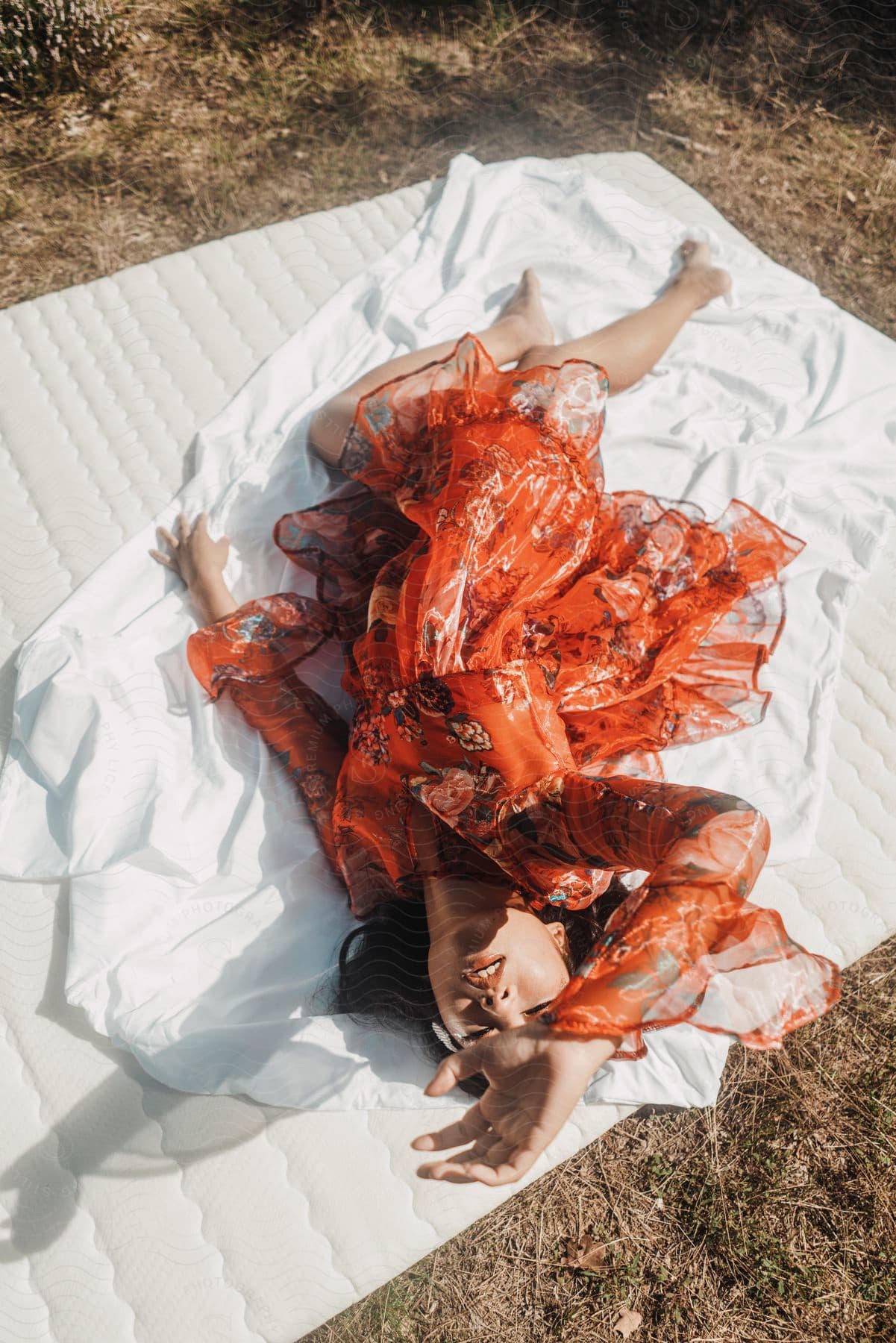 A woman wearing a red print dress is lying on a white blanket on the ground as she raises her hand in over her head