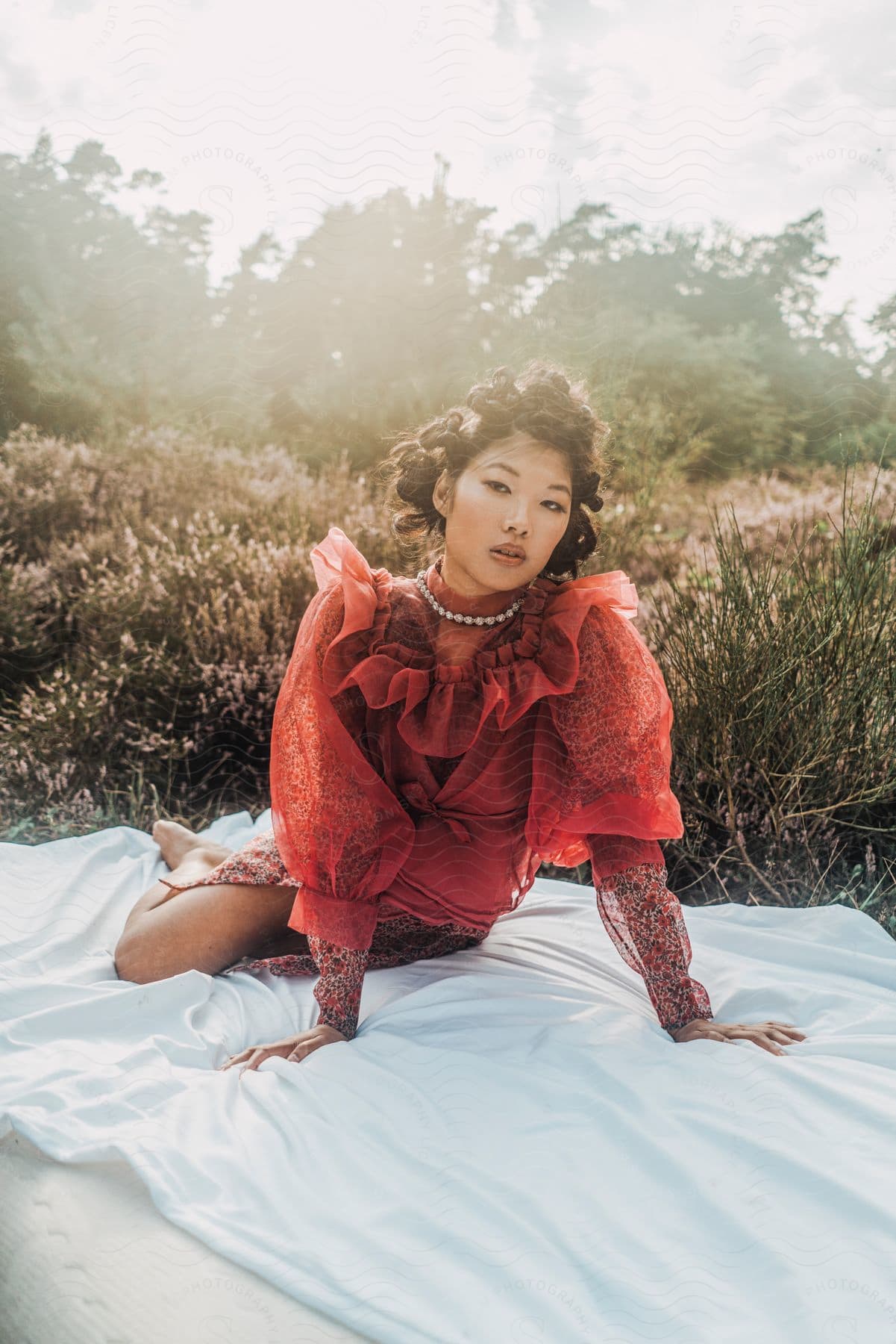 An asian women is sitting on a blanket in a meadow in a red dress.