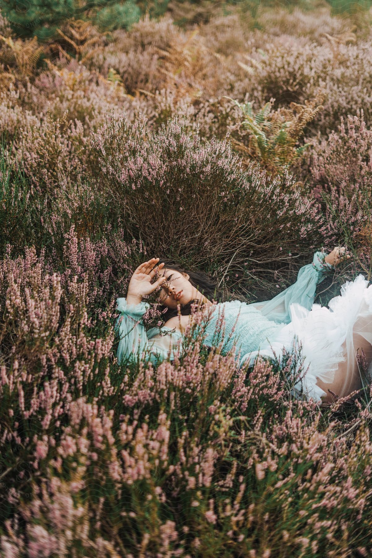 Sun shines upon a woman lying in a field of plants and flowers as she holds her hand in front of her face