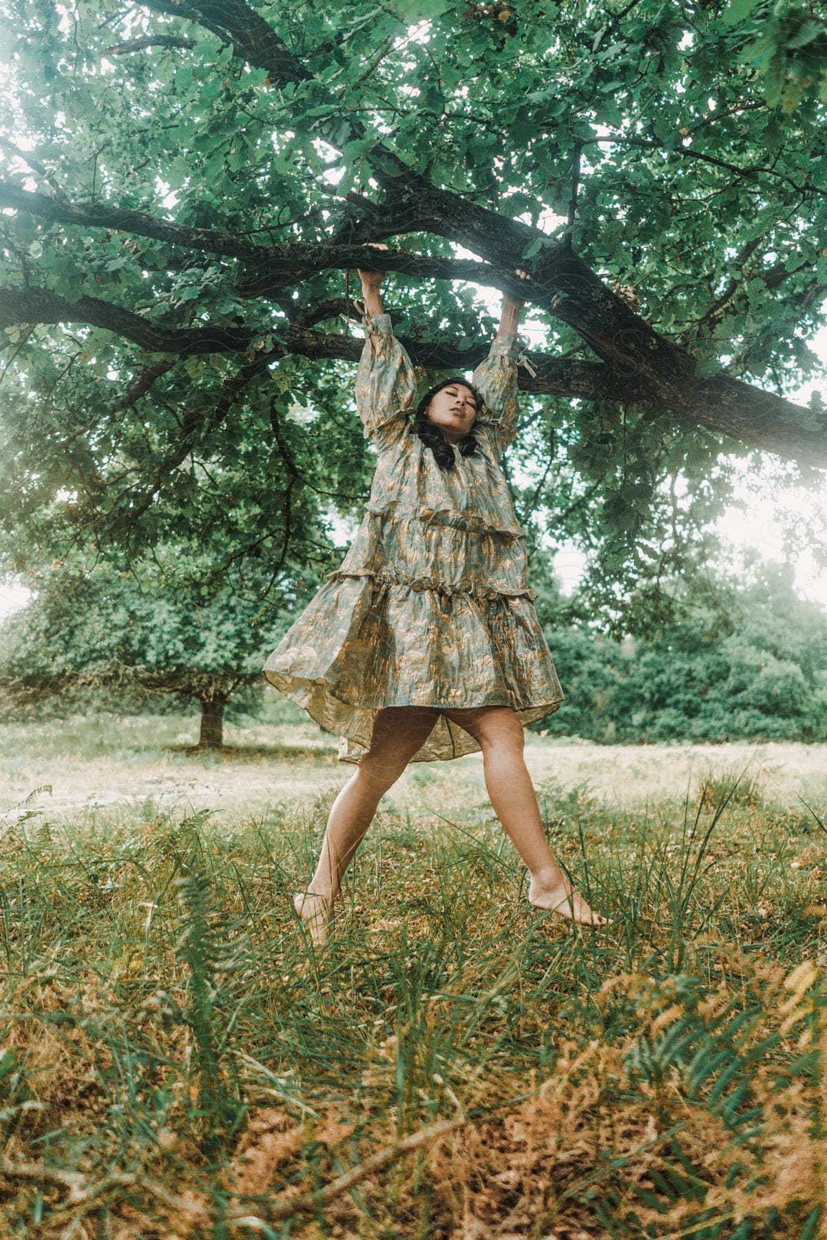 A barefoot woman wearing a dress hanging from the branch of a tree in the park