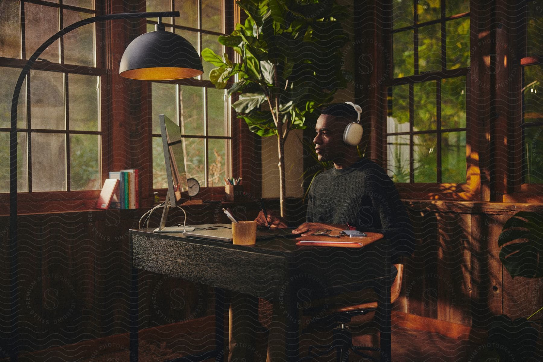 A man wearing headphones is sitting at a desk working on a computer in a room filled with windows