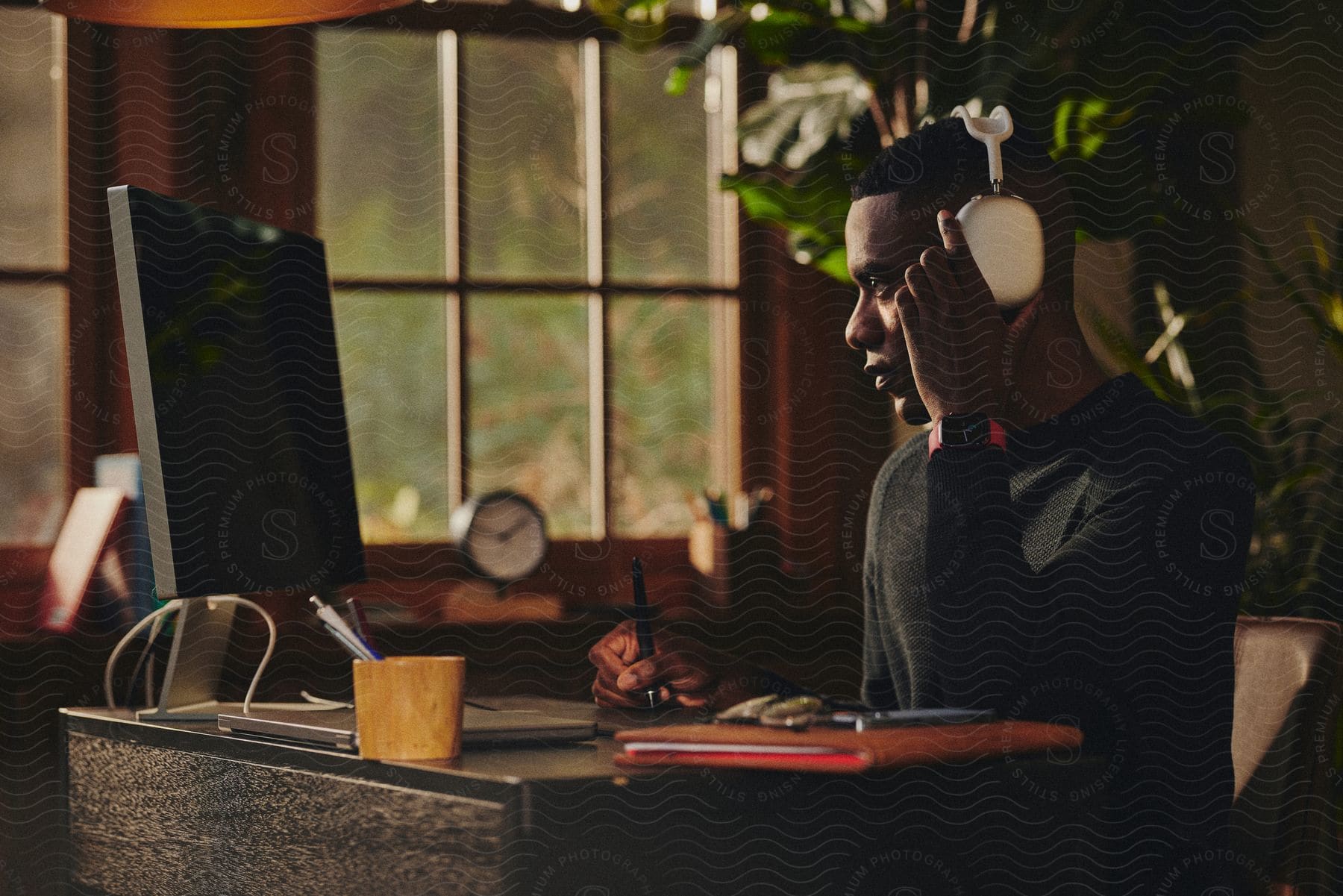 A man in a home office, wearing AirPods Max and writing in a notebook, with a Apple mac desktop and pens on his desk.