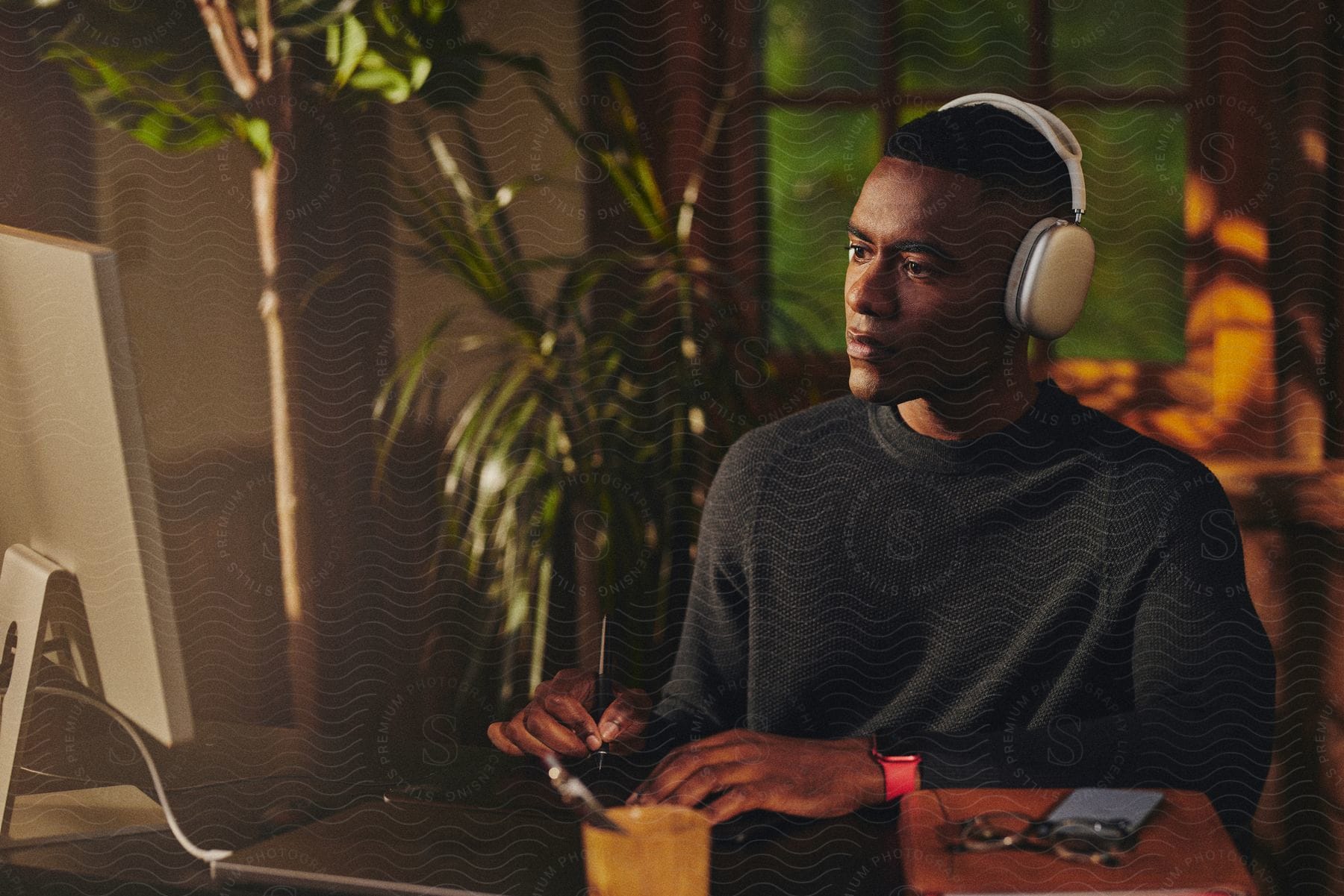 A man wearing headphones is sitting at a desk holding a pen as he looks at the computer monitor