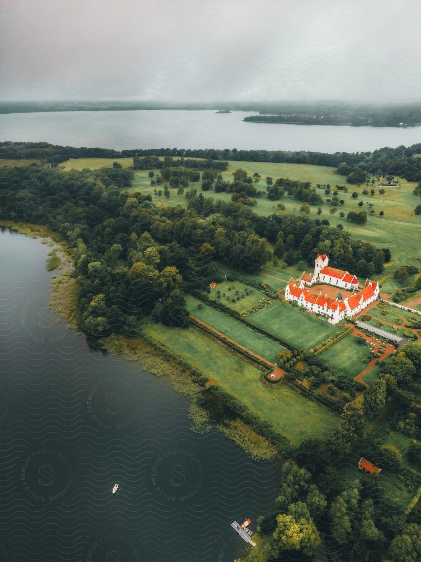 Red roofed historical buildings contrast with fields and nearby woods a short distance from lake on an overcast day.