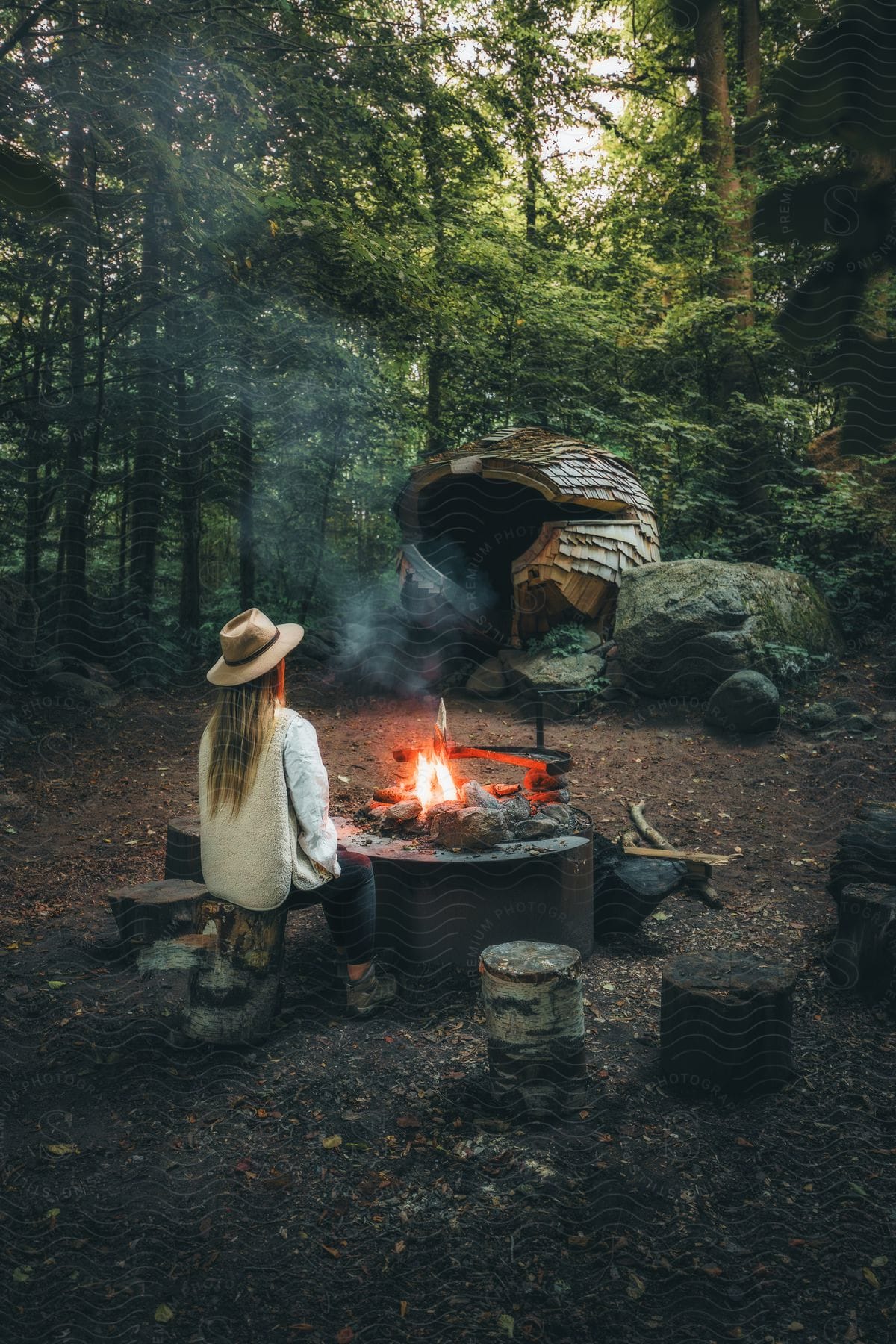 A woman with a sunhat, wearing a vest and white shirt, sits by a campfire with a wooden tent in the background, in the forest.