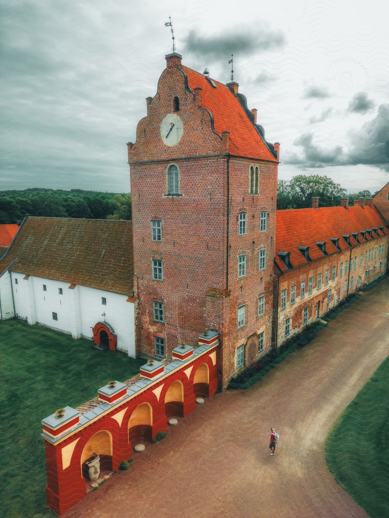 Bäckaskog Slott, a brick castle with an orange roof and clock tower, stands alone on a cloudy day, with a lone tourist walking on the dirt road in front of it.
