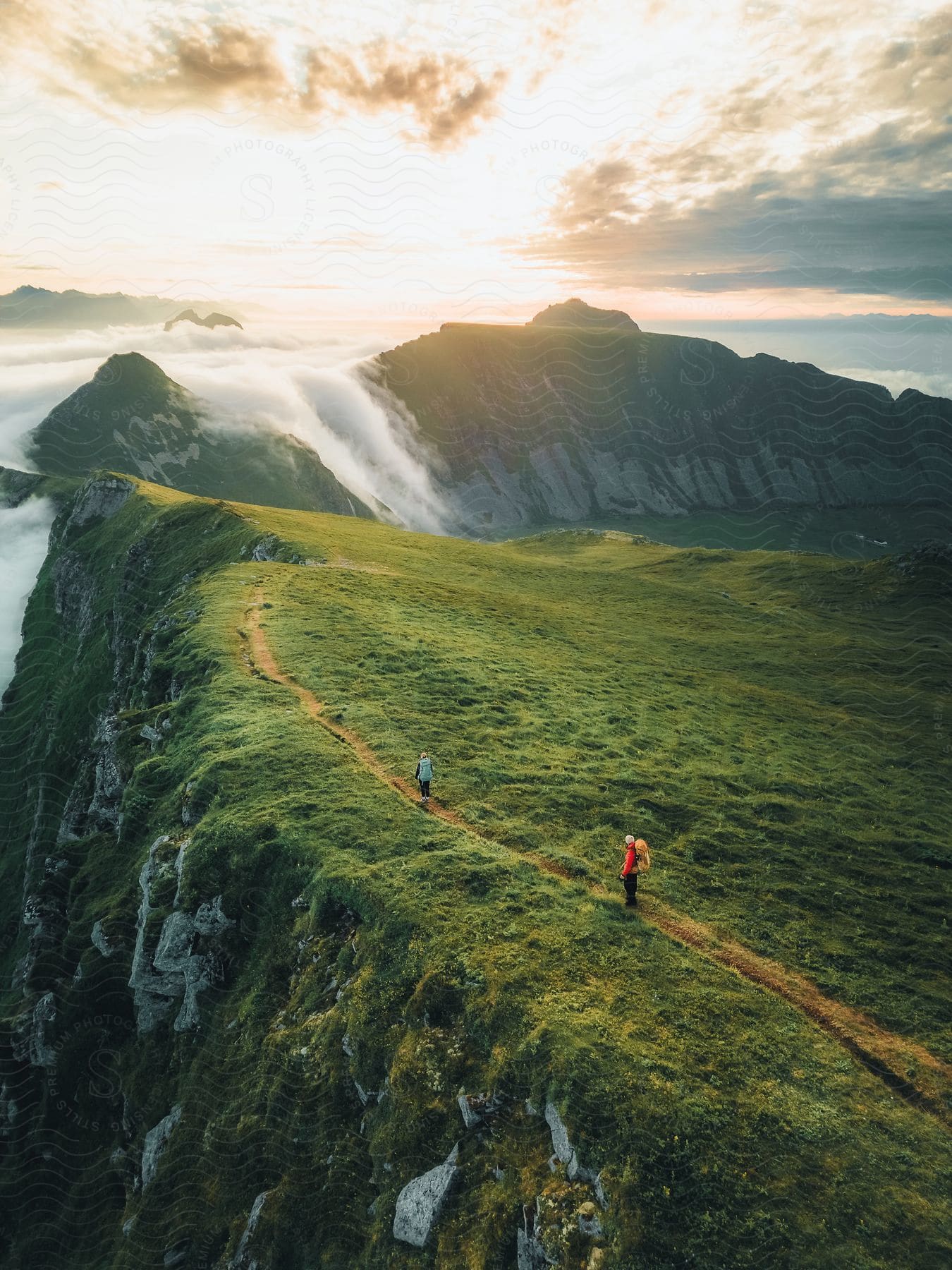 Two hikers traverse a lush green hillside cliff at sunset, as a low-hanging cloud rolls over the mountain range beyond.