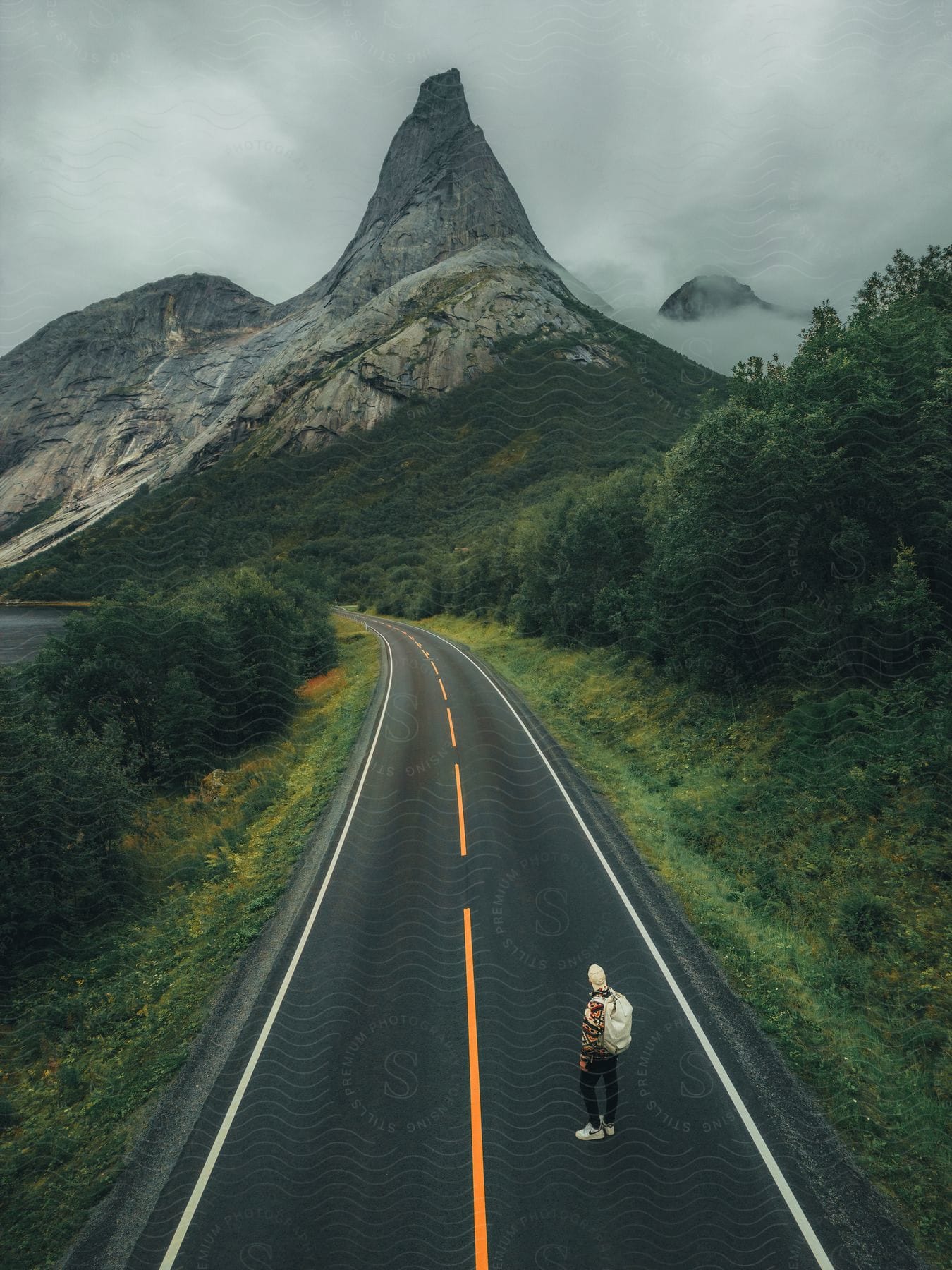 Hiker stands on highway curving through woods and toward foggy mountains on an overcast day.