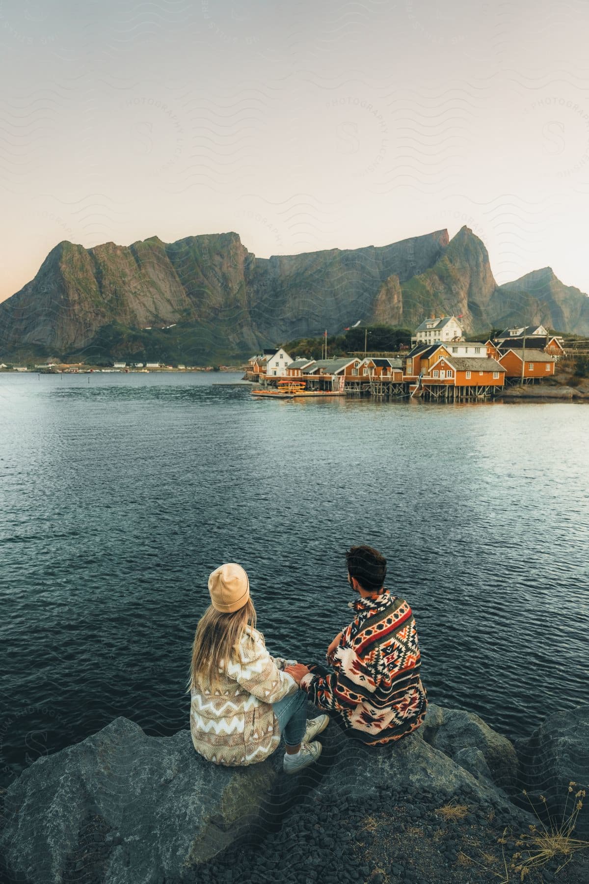 A man and woman hold hands while sitting on a boulder and overlooking a small coastal village.