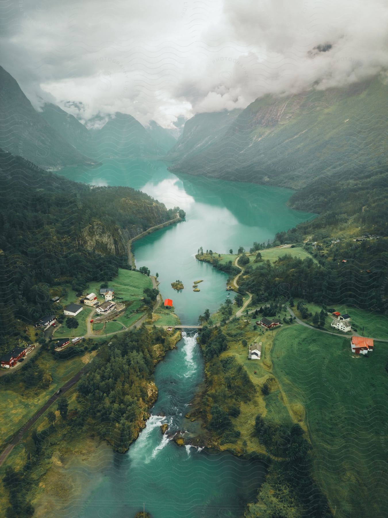 Aerial view of a lake surrounded by mountains and a small rural town, divided by a river and connected by a bridge, under a cloudy sky.