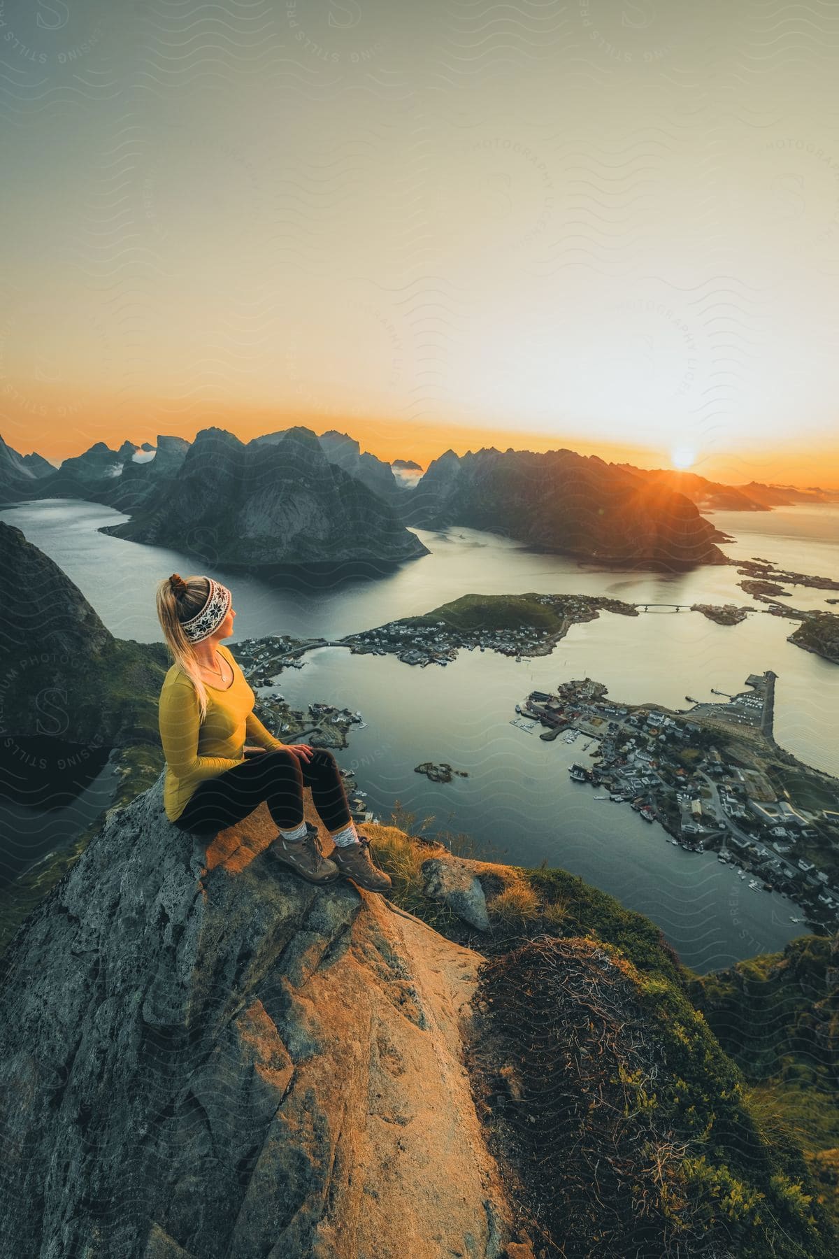 A Female Hiker Sits On Top Of A Mountain Peak At Sunset While Overlooking The Coastal Community Below