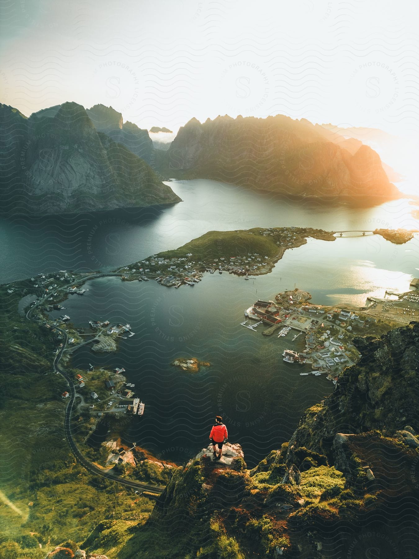 A person in an orange jacket stands on a hill cliff overlooking a coastal town, mountains, a body of water, and mountain islands, with a road leading into town, docks with boats, and piers at sunset.