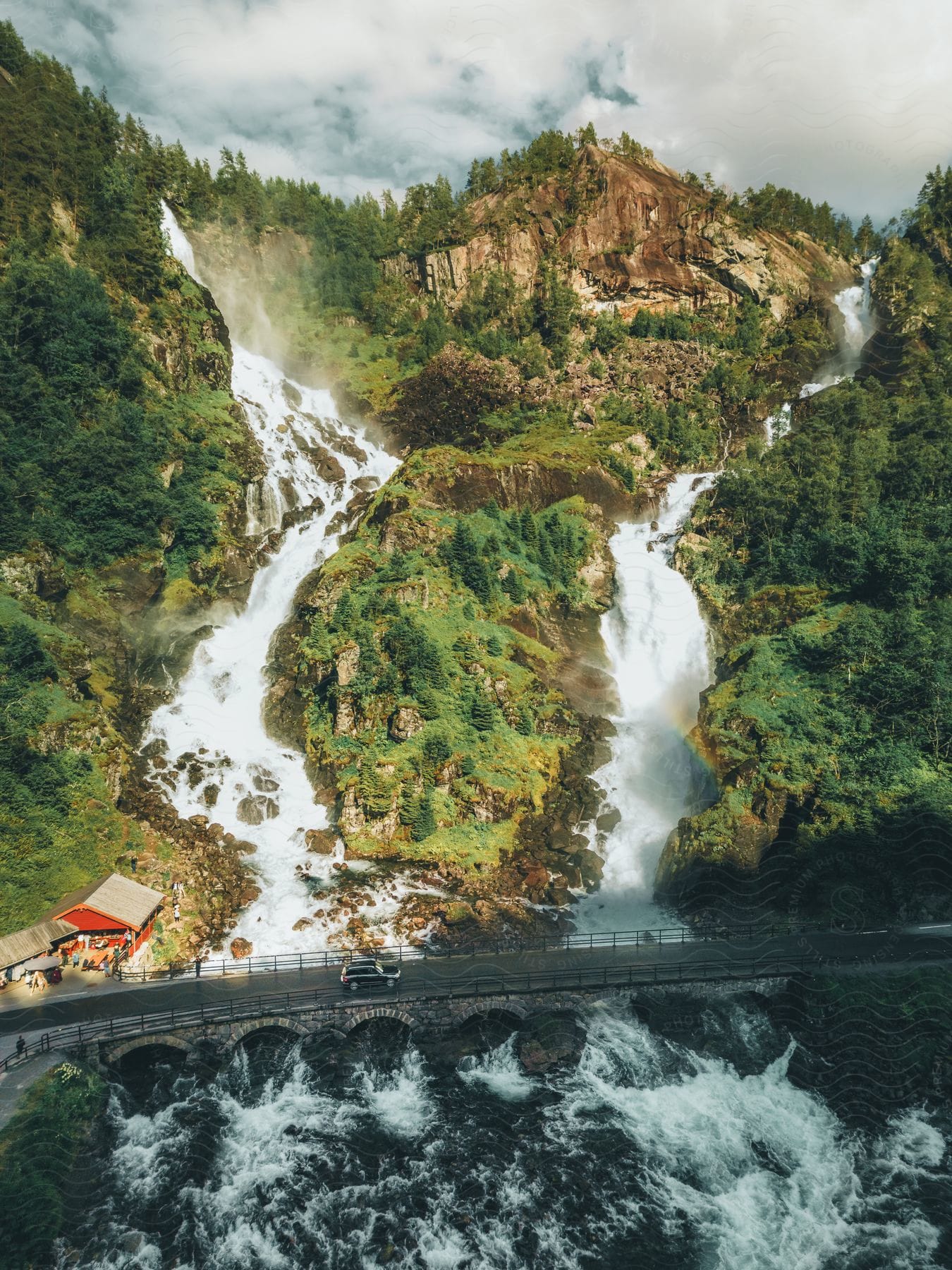 A scenic duo waterfall flows down a rocky hillside into a river that merges under a bridge, with red shops on the lower left and people and a black SUV crossing the bridge.