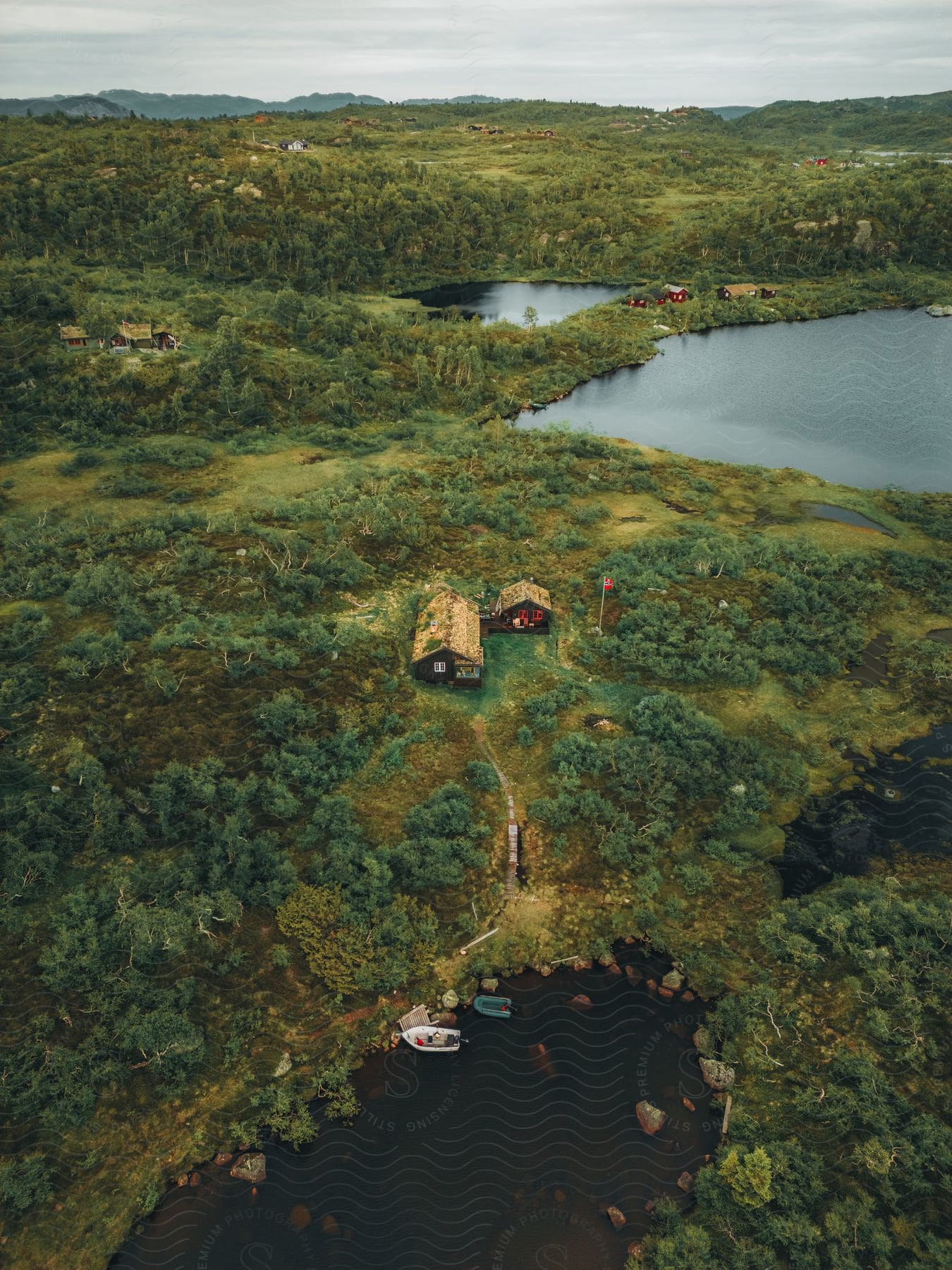 A small cabin on a hill overlooking a forest-surrounded lake, with boats on the shore and other houses in the distance.