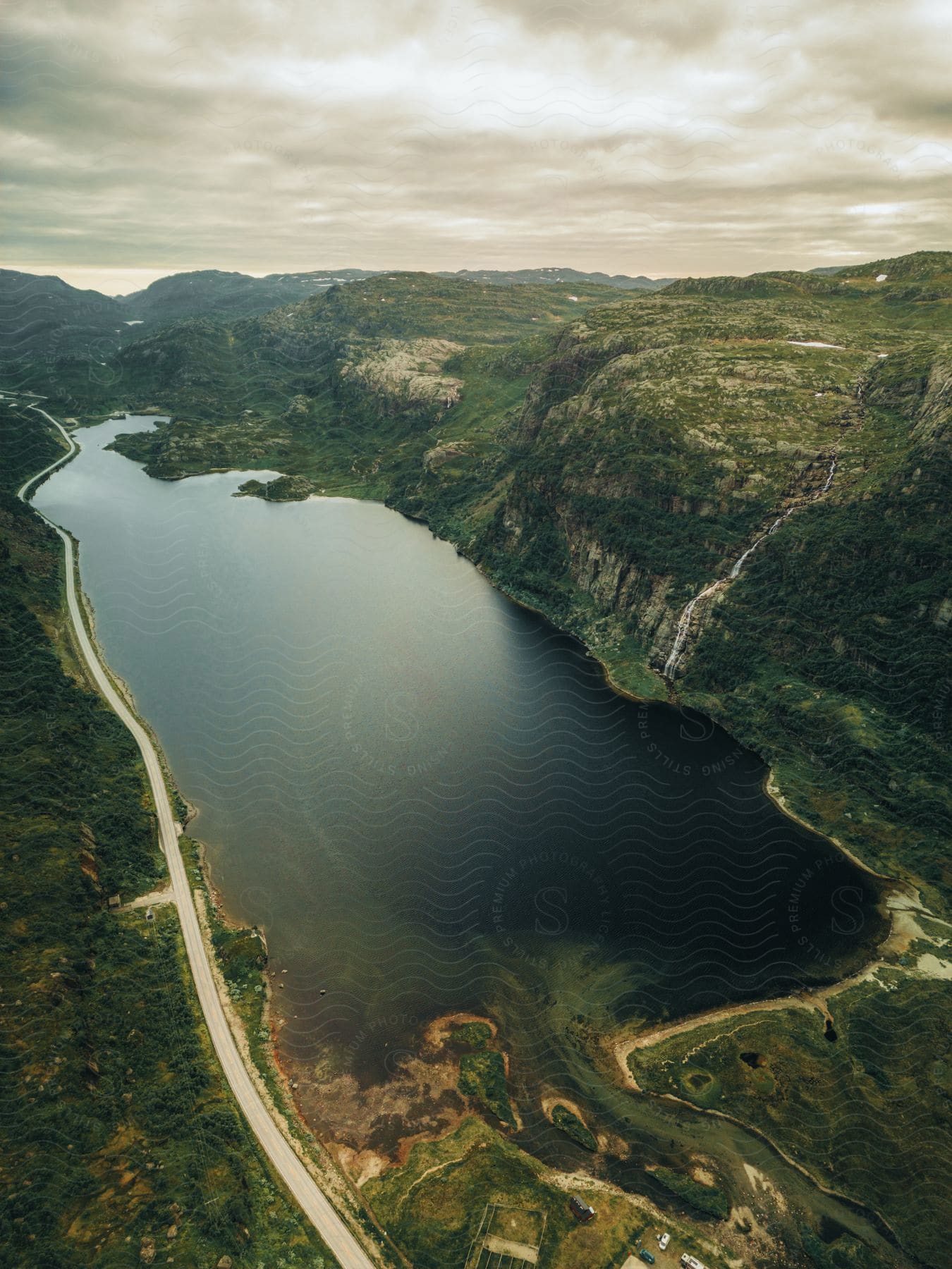 A road runs alongside a lake and a steep mountain canyon under a cloudy sky