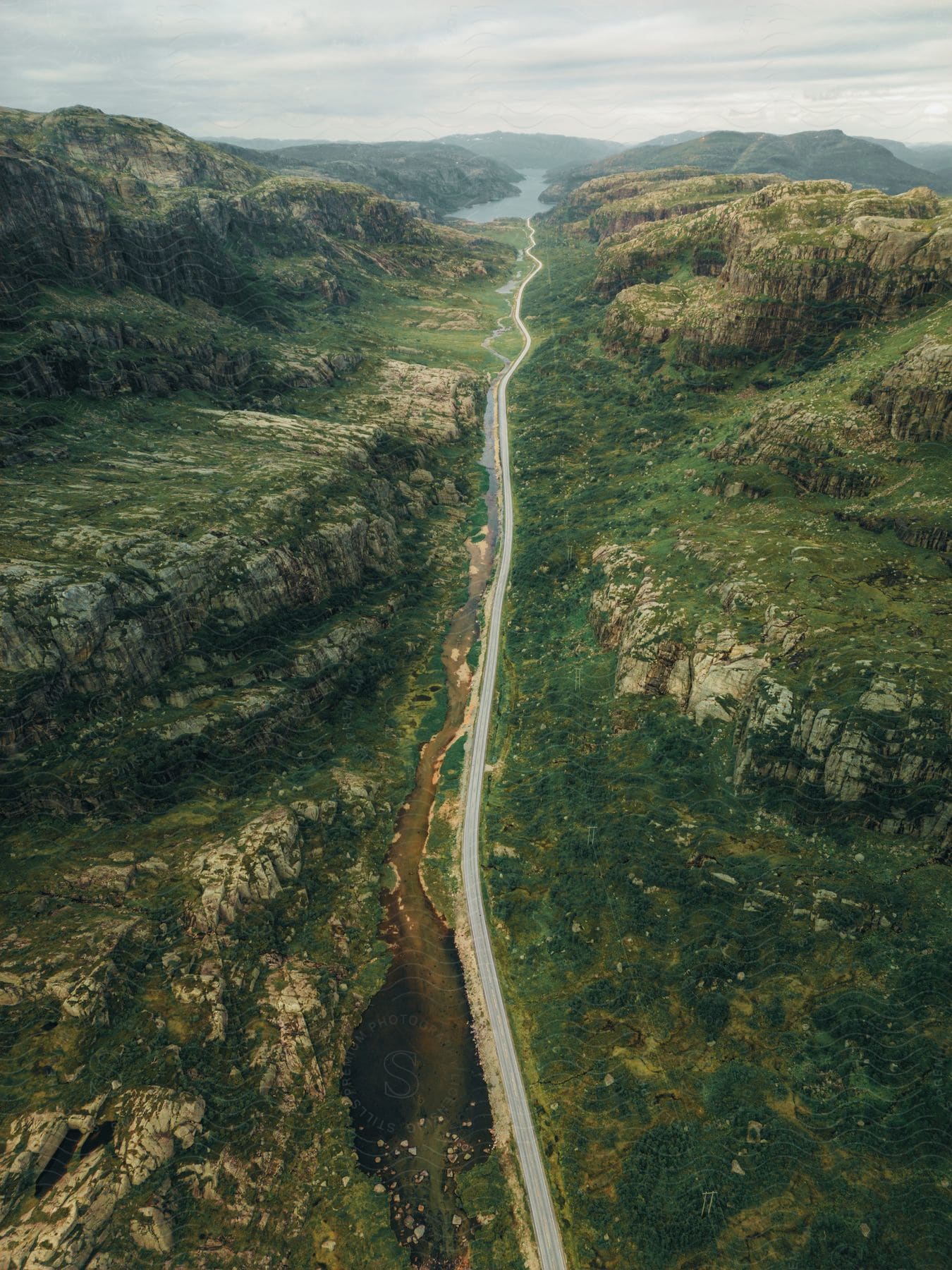 A stream runs along a long empty road through a mountain valley under a cloudy sky