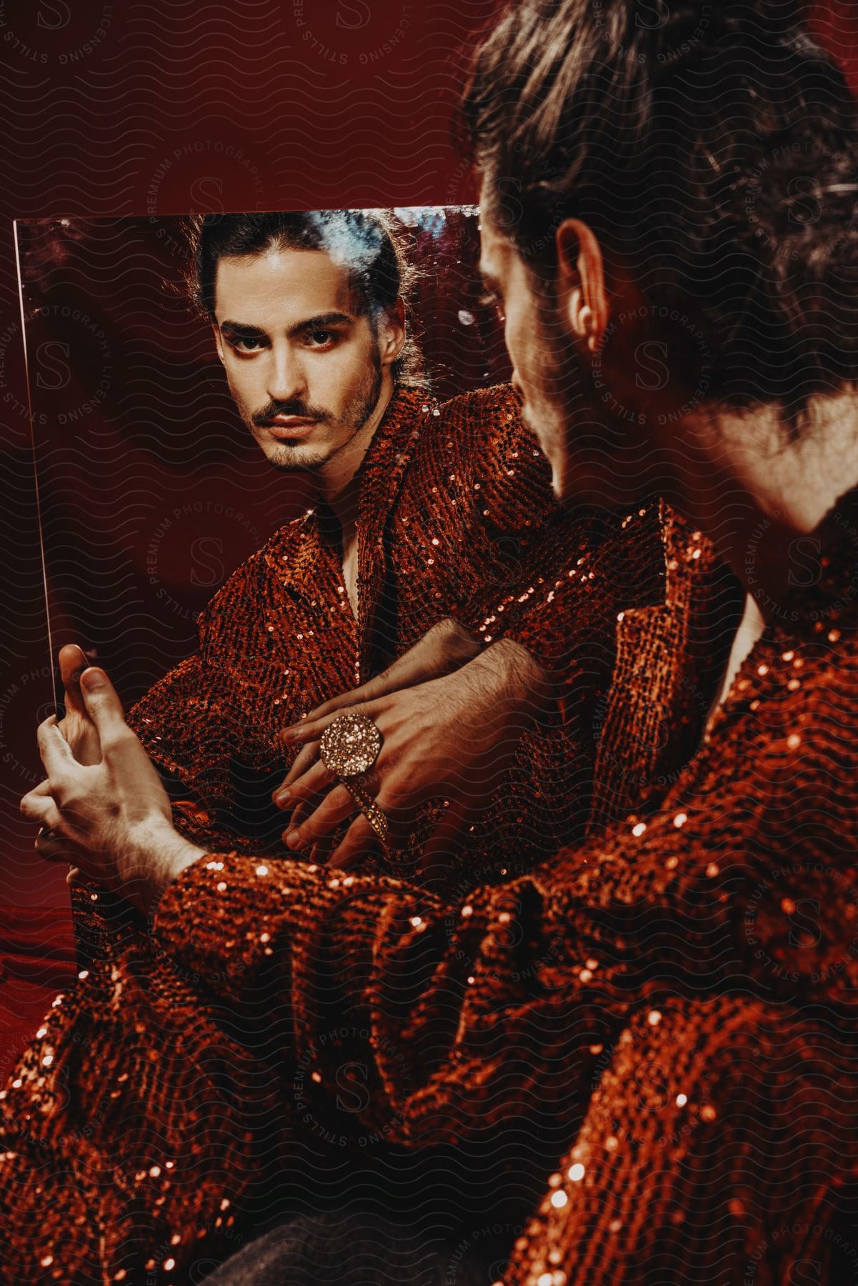 A red sequin-jacketed man sits on a black cushion in a red studio, holding a large mirror and gazing at his reflection, trying to touch it while wearing a massive diamond ring.