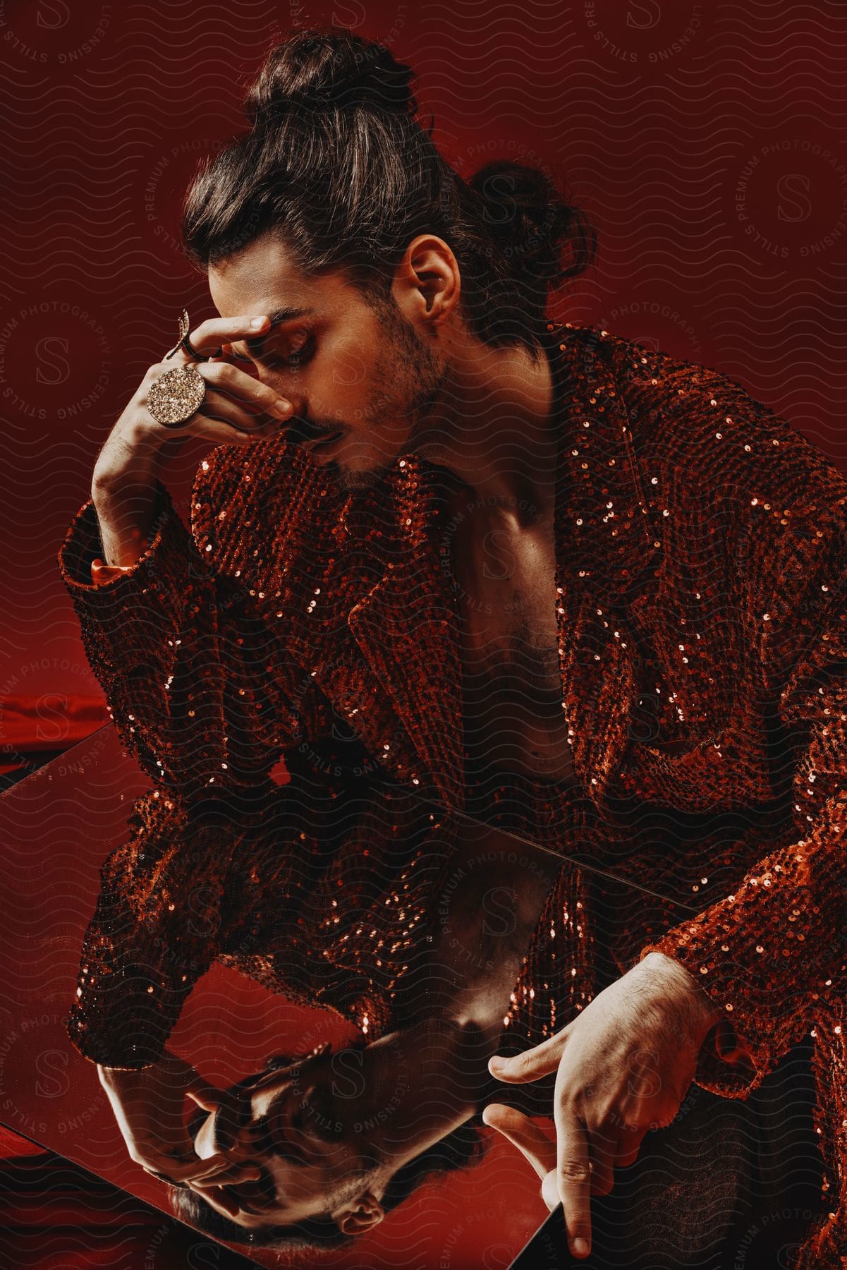 A shirtless man in a red sequined jacket sits on a black cushion, holding a large mirror in his lap and gazing at his reflection, a massive diamond ring on his hand and one hand on his face.