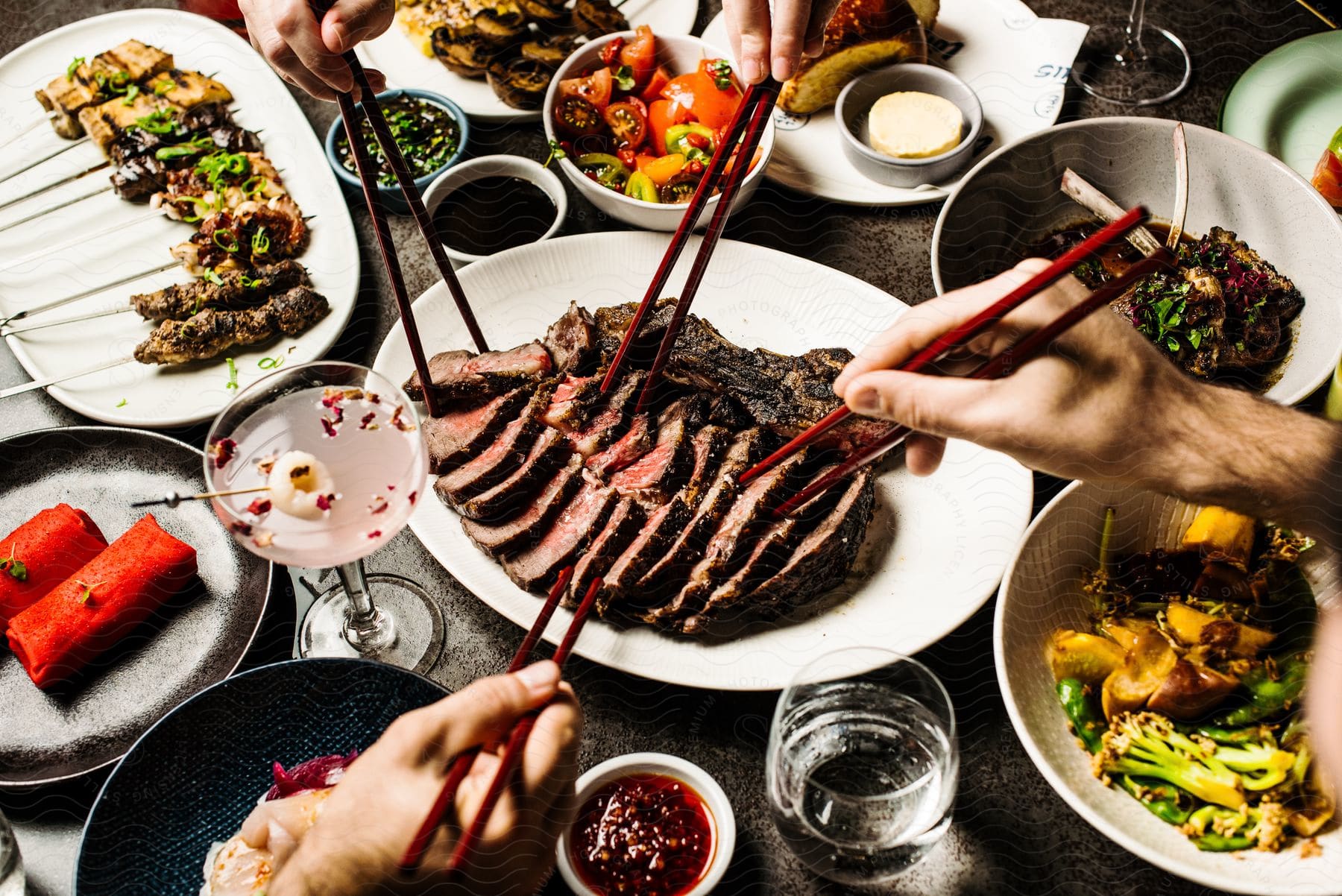 A table set with Asian food and drinks, with four diners using chopsticks to dig into a sliced beef dish in the center.