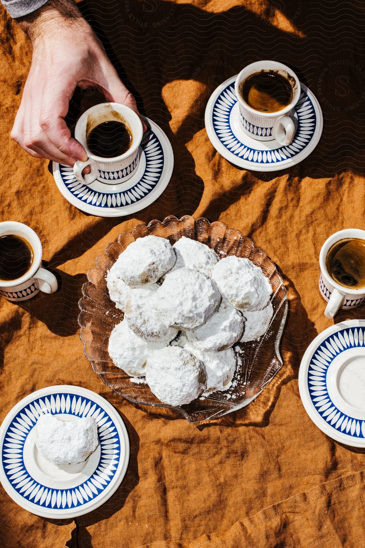 A man is holding a cup of coffee with many other cups situated around a plated dessert.