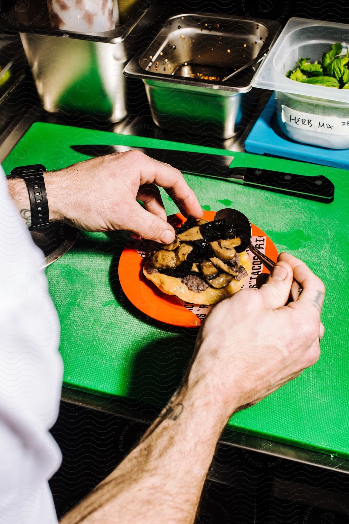 A chef is plating a taco with sauteed mushrooms and beef strips on a corn tortilla, with other food items in the background.