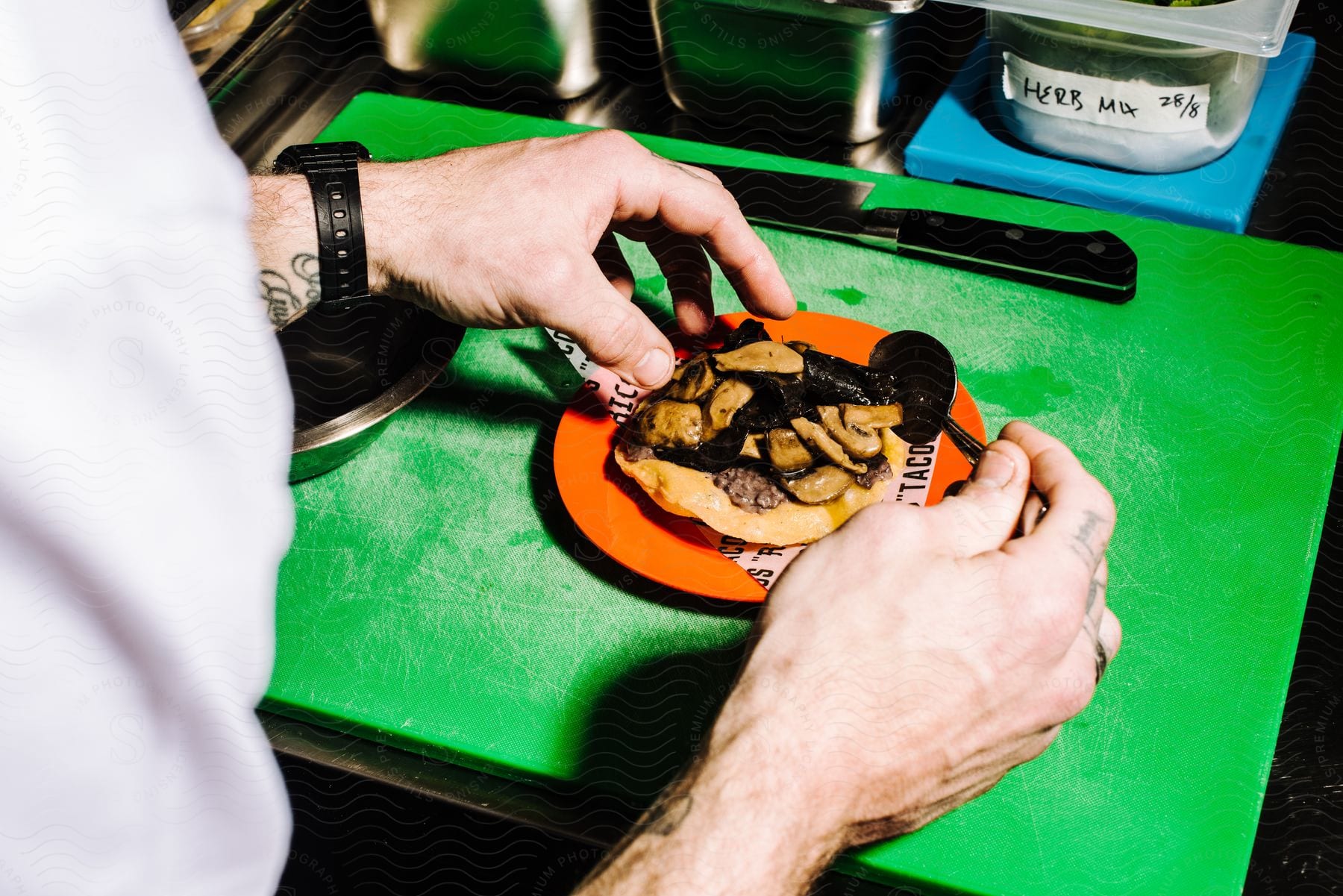 A chef in a restaurant places sautéed mushrooms with beef strips on a corn tortilla taco on an orange plate on a green cutting board.