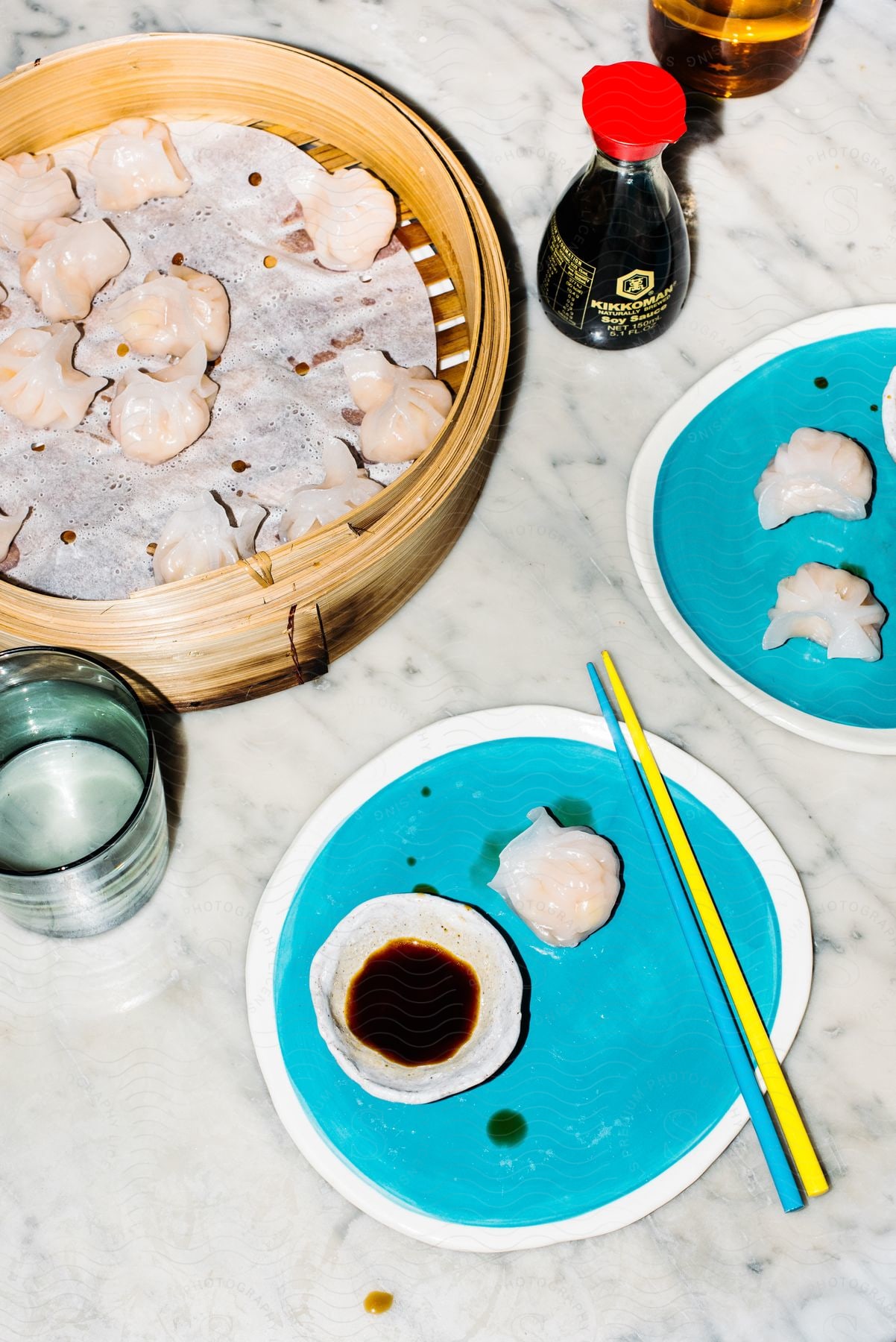A marble table with blue and white plates of shu-mai dumplings, yellow and blue chopsticks, a bottle of soy sauce, a Chinese steam basket with more dumplings, and a glass of water.