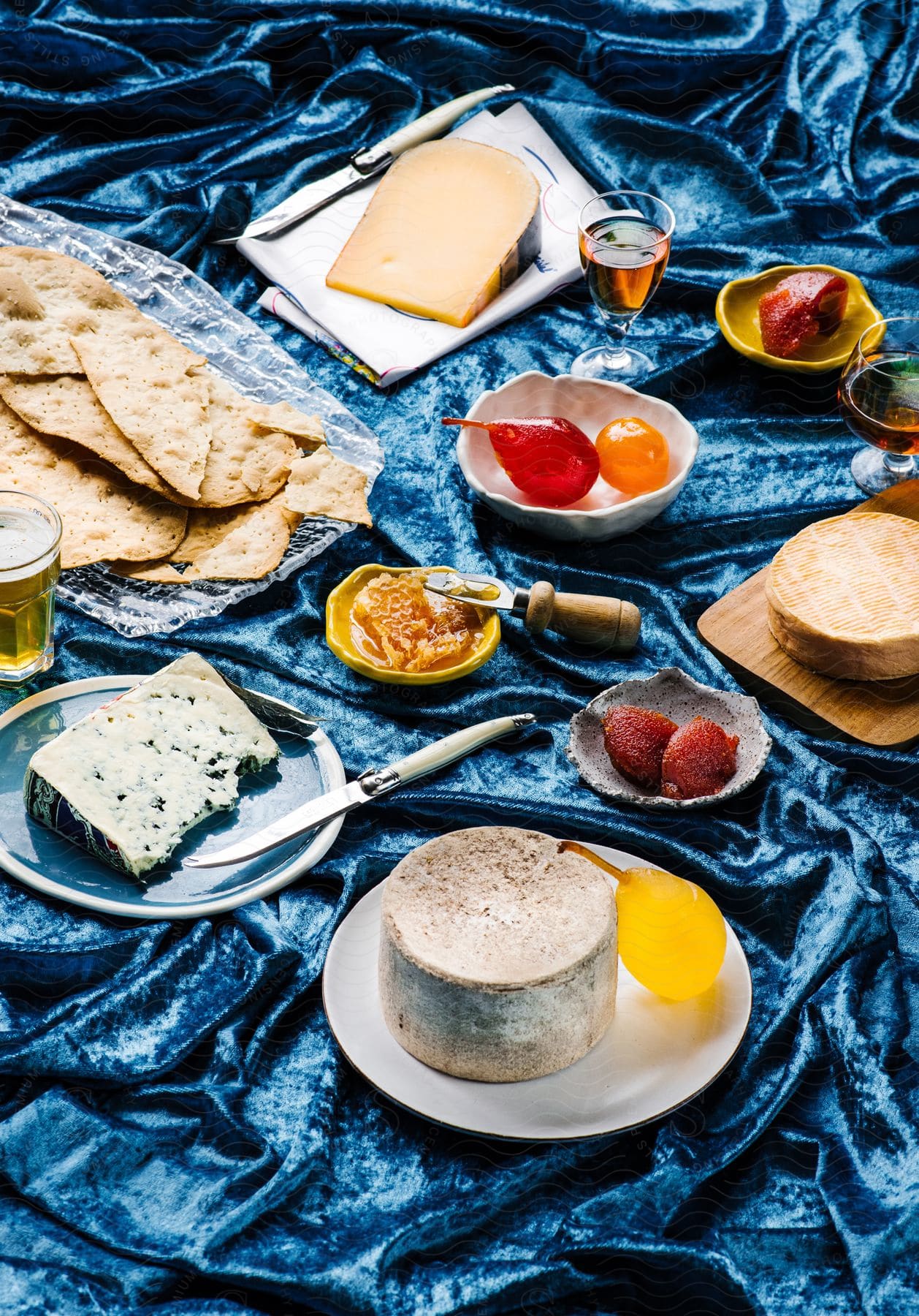 A bountiful blue velvet blanket spread with plates full of food, drinks, cheese, fruits, nan bread and cheeseboards, accompanied by knives and a variety of alcoholic drinks in various glasses.