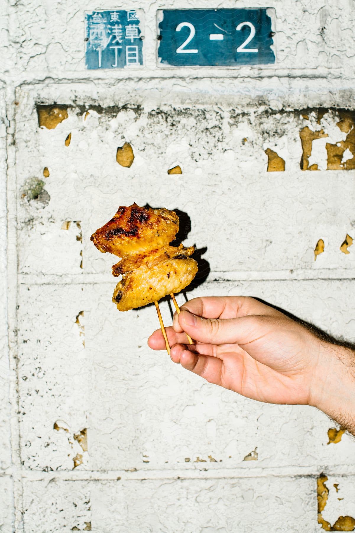 A hand holds grilled chicken yakitori in front of a wall with a "2 - 2" sign near a chipping building.