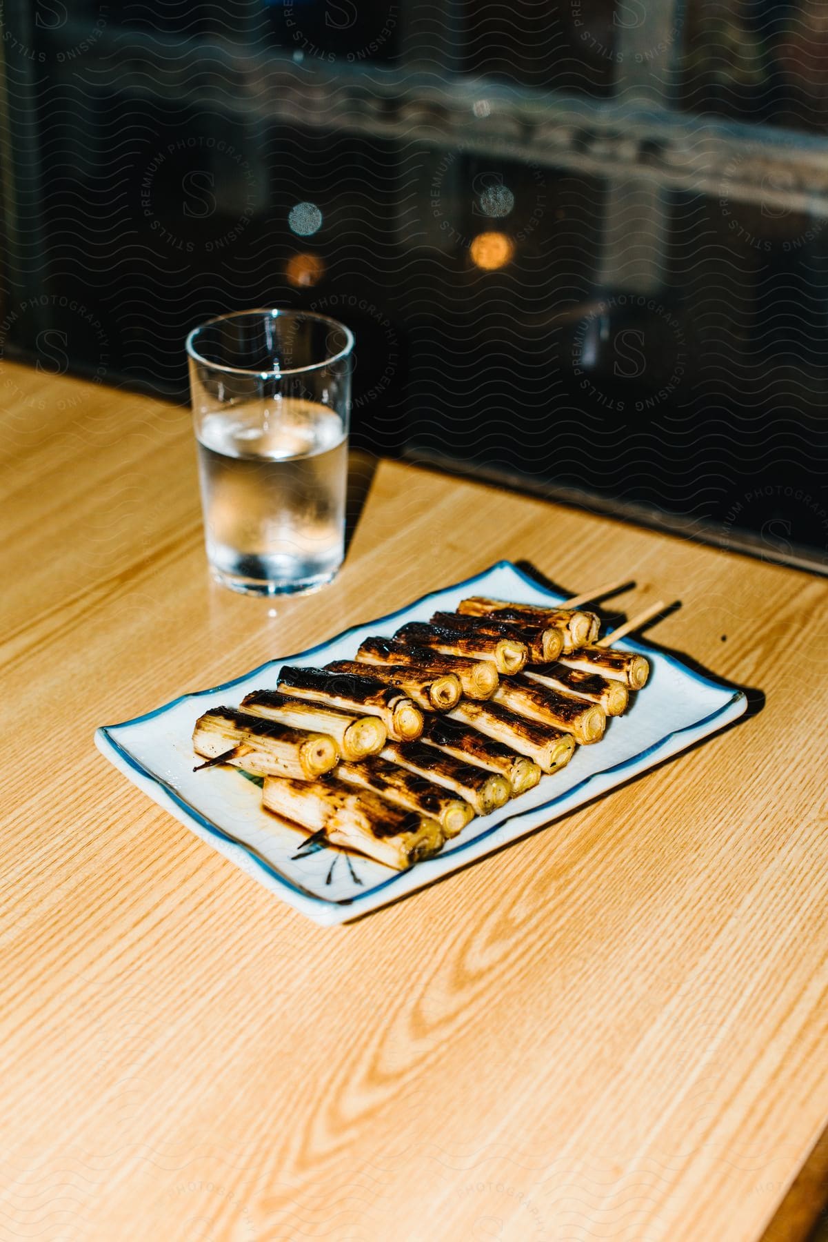 Skewered food and beverage sit on table near window at night.