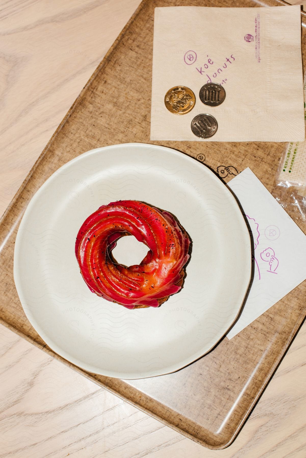 A red frosted donut on a white plate sitting on a placemat with a drawing underneath and writing on a napkin