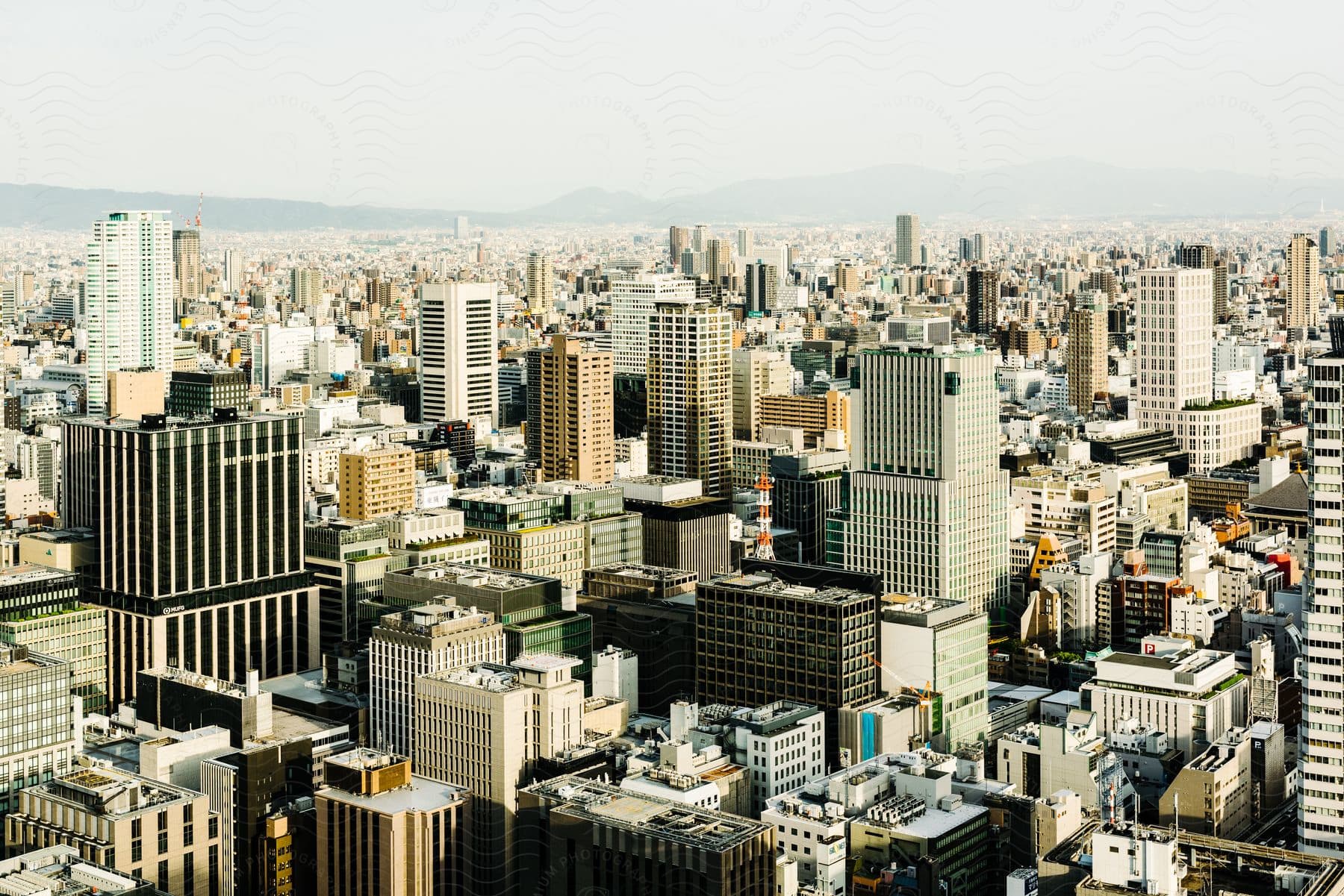 An aerial view of a major Japanese city on a hazy day, with tall buildings, mountains in the background, and an endless view of rooftops.