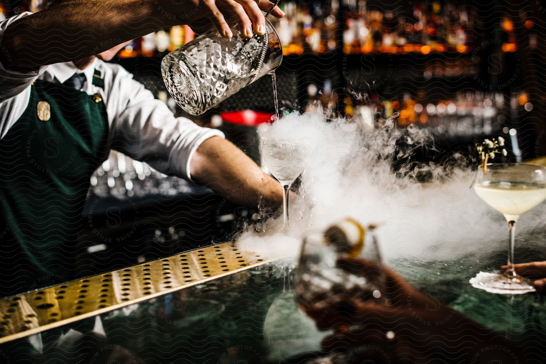 A skilled bartender behind the bar pours a martini into a glass with dry ice, creating a smoky spectacle.