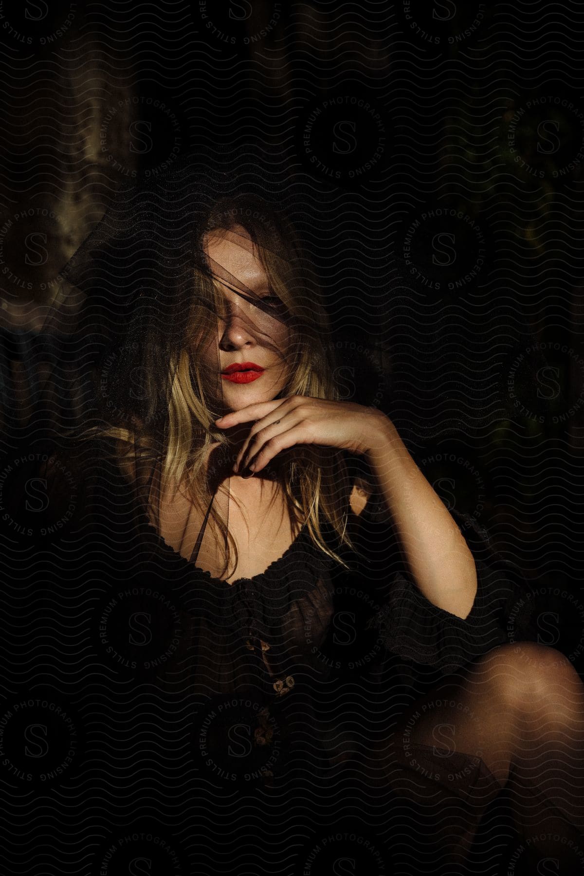 A blonde in sheer black with a mysterious veil and vivid lips sits in a dimly lit room.