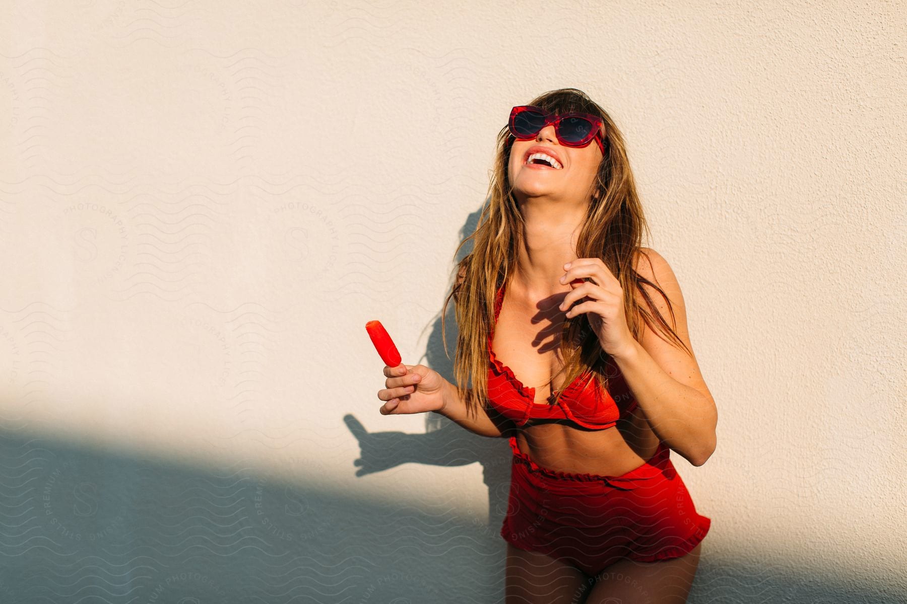 A woman wearing a red bathing suit holding a popsicle has her head back laughing