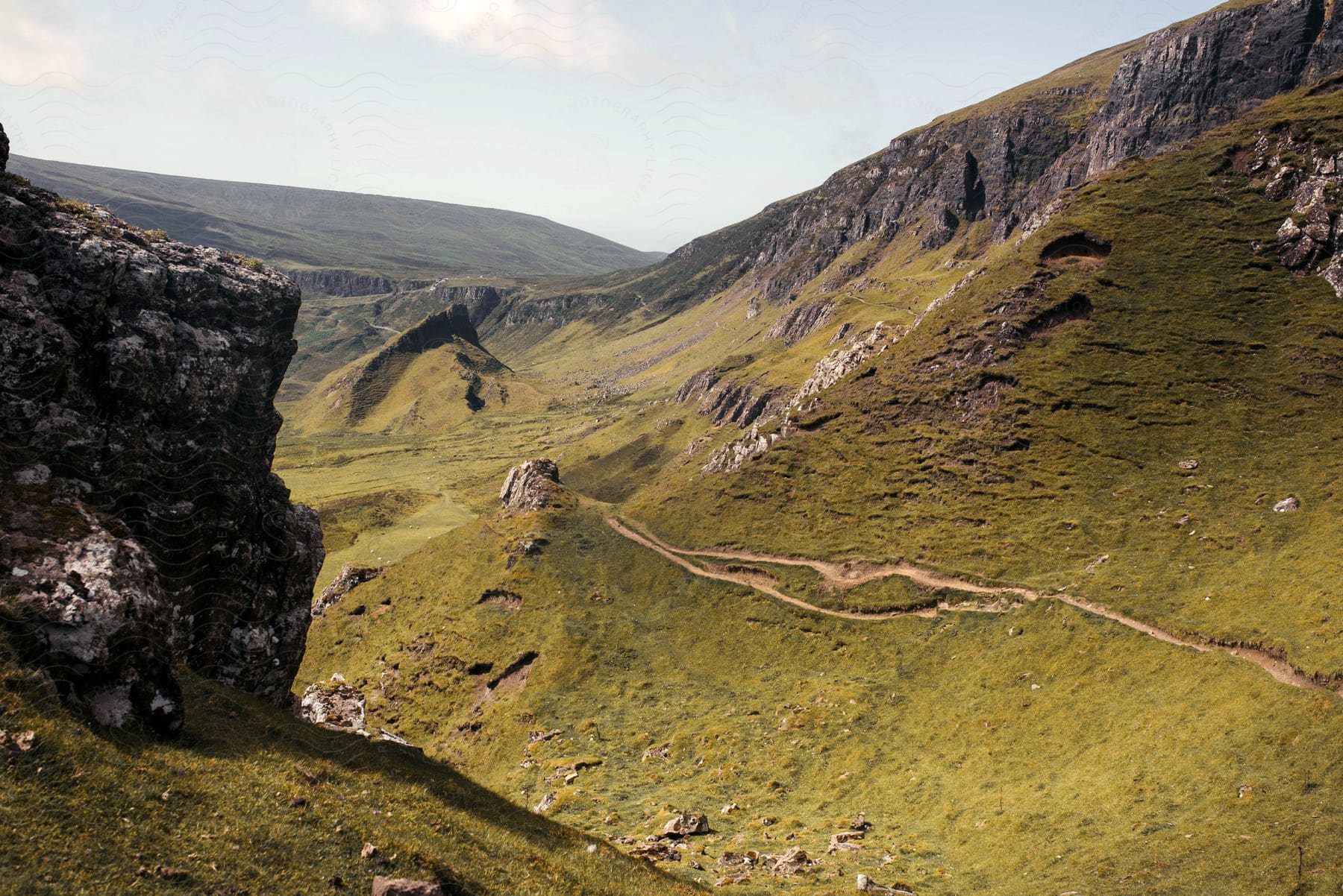 A road runs along the side of a steep mountain overlooking the valley below with mountains in the distance