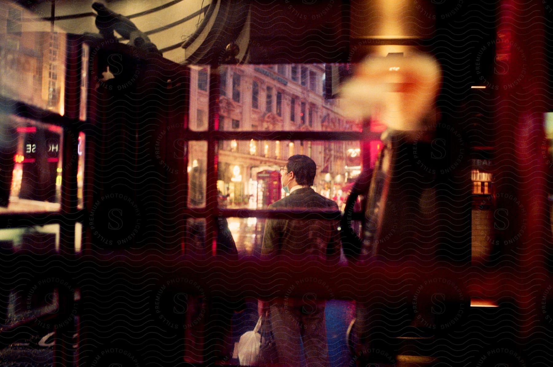 A man stands in a bustling London street, viewed through a phone booth's window, with an illuminated old-style hotel ahead.