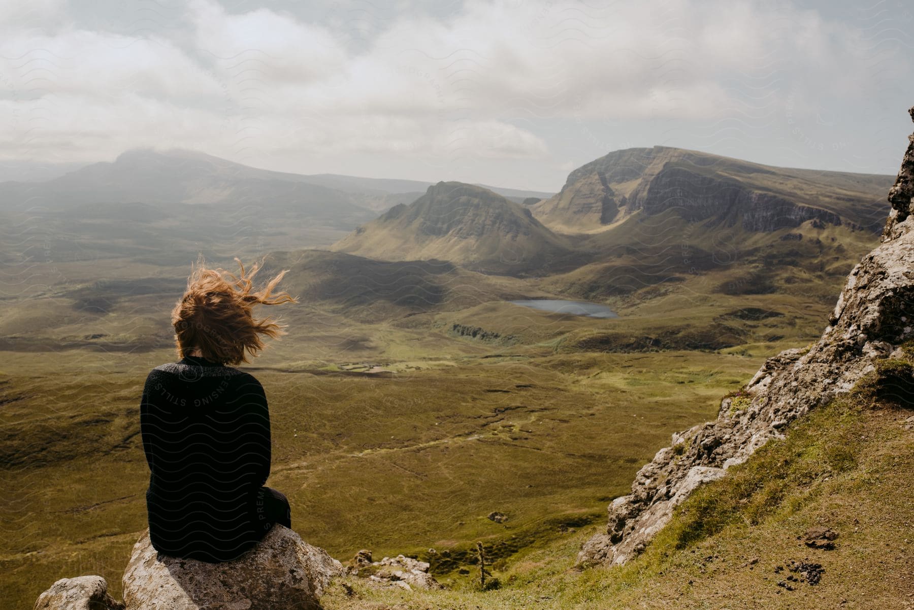 A woman sits on a rocky ledge along a mountainside looking at mountains in the distance as her hair blows in the wind