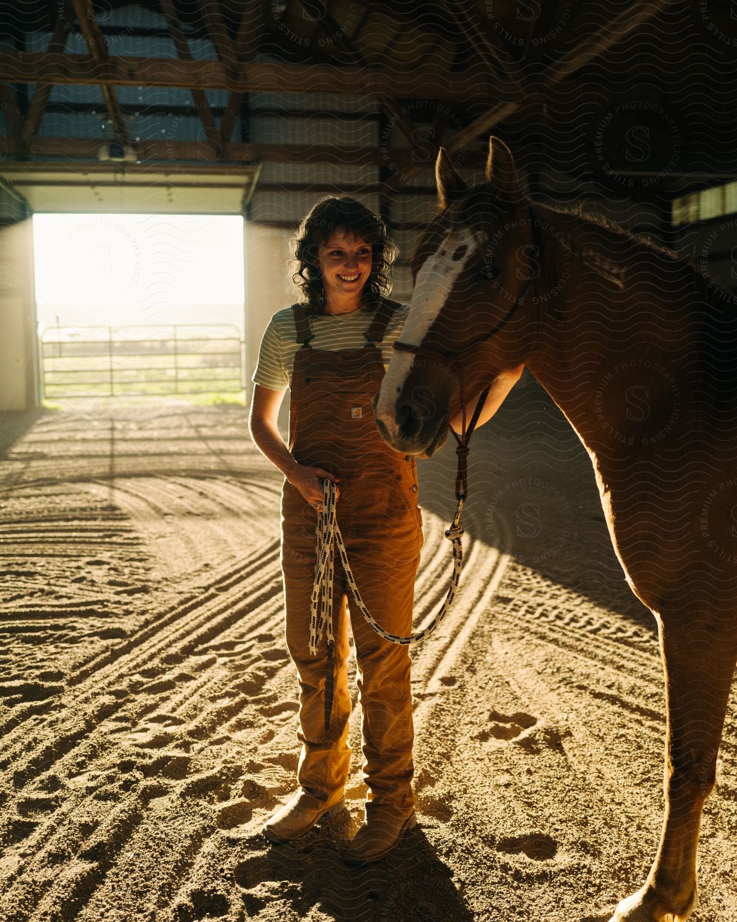 Stock photo of a woman in overalls standing in a large stable smiling and petting a horse