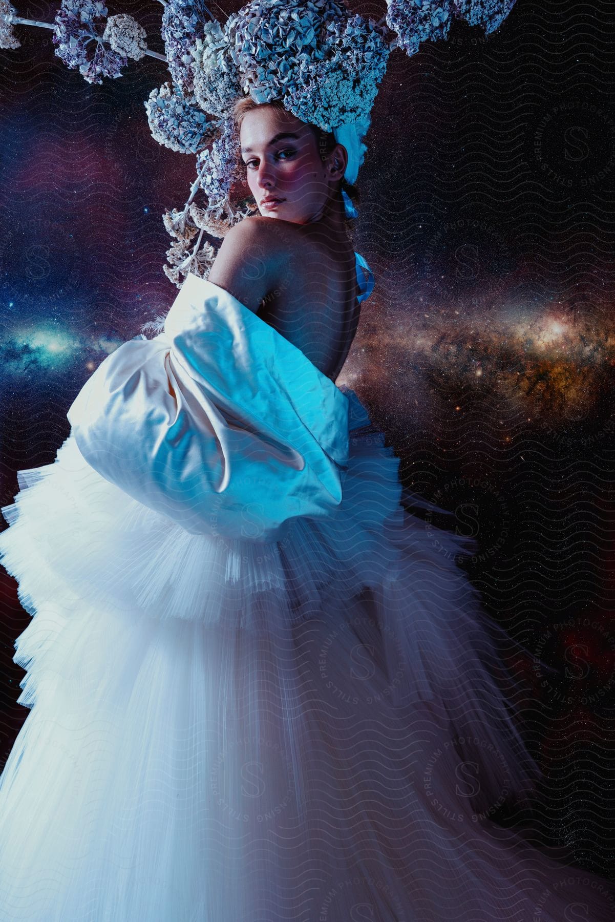 Stock photo of wearing a long gown, a lady puts on flowery accessory hat that resembles part aluminum foil and part shrubbery on a galaxy background.