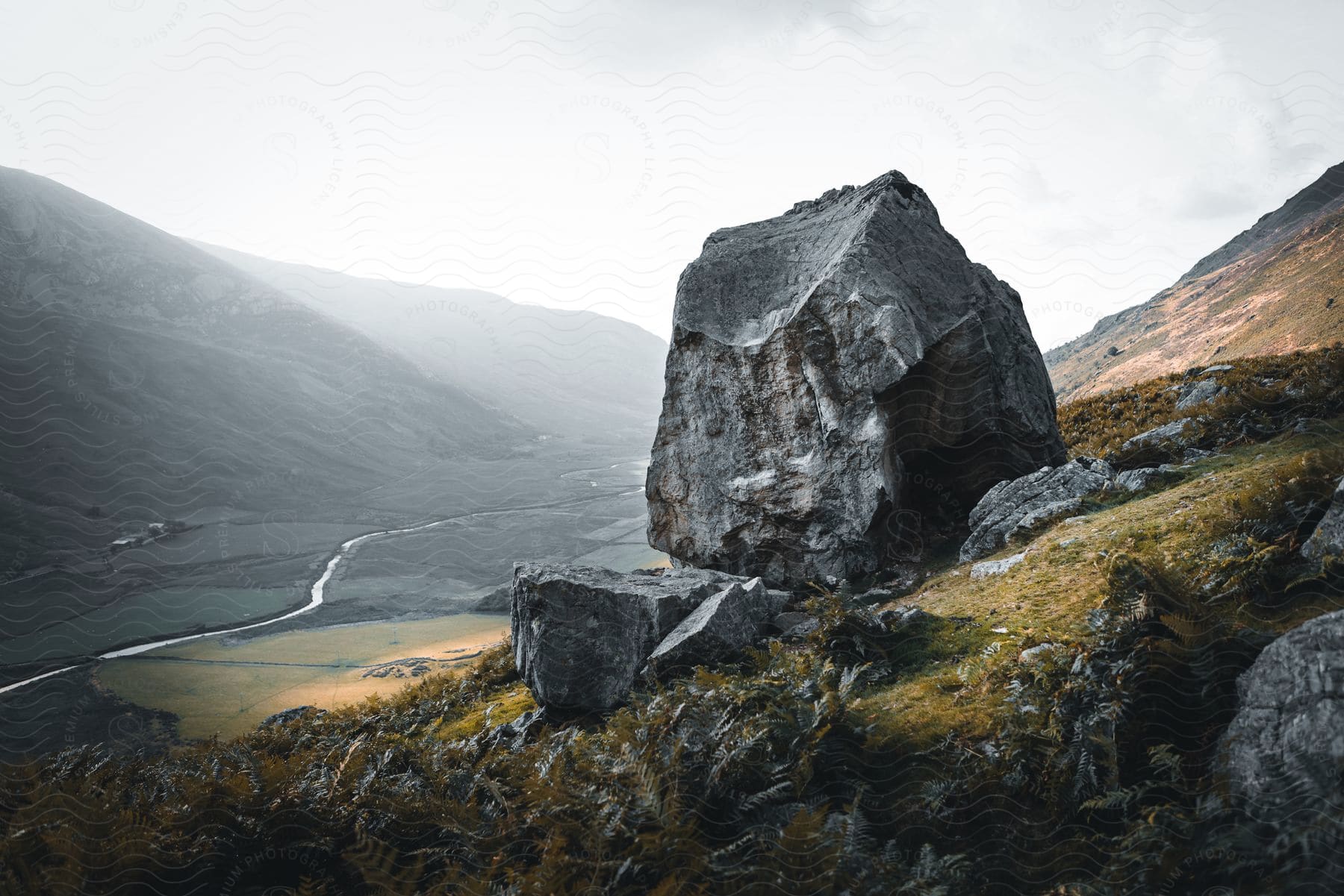 A large boulder on the side of a mountain with a river winding through a valley in the background.