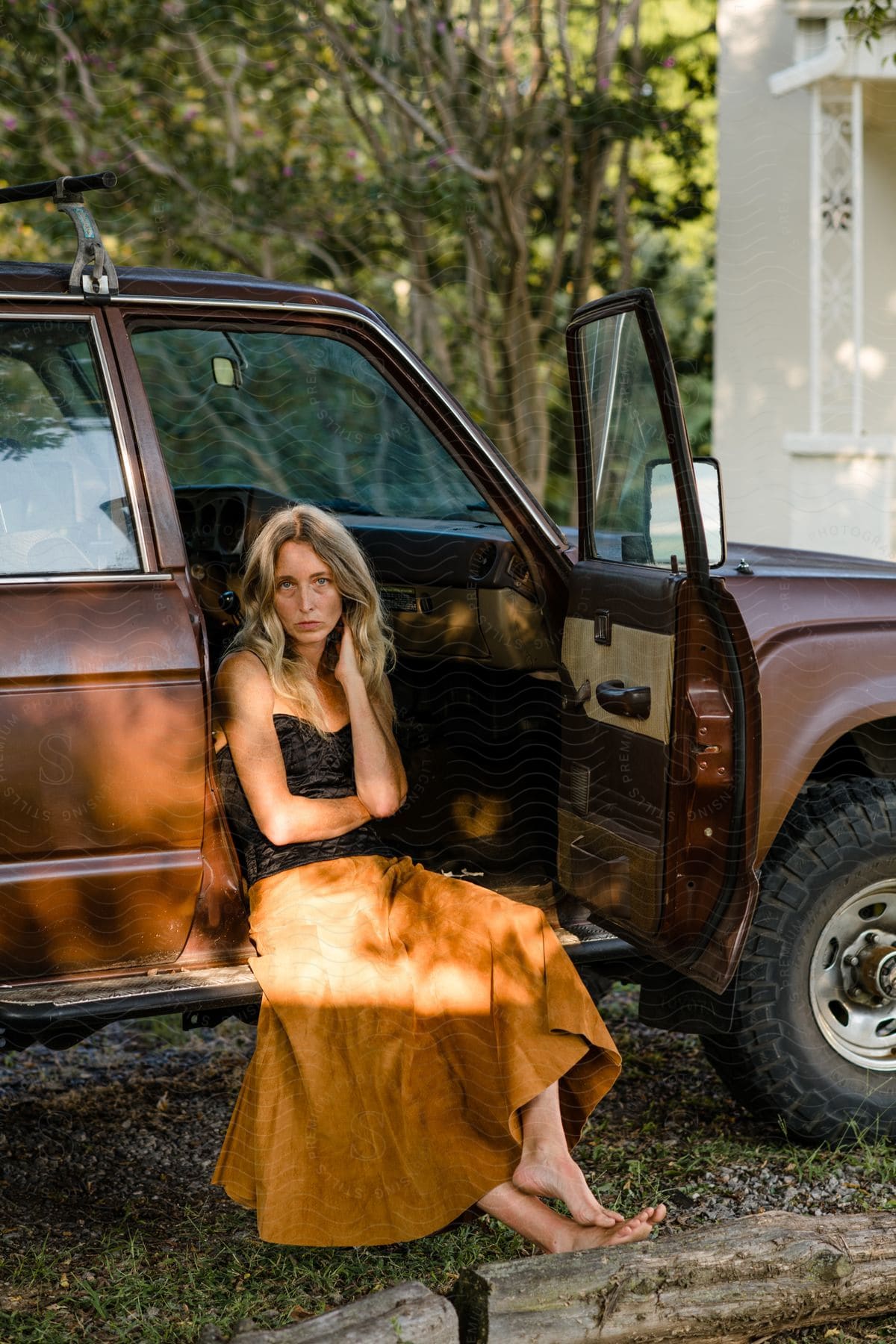 A woman with long blonde hair and wearing a long skirt is sitting inside the open door of a jeep