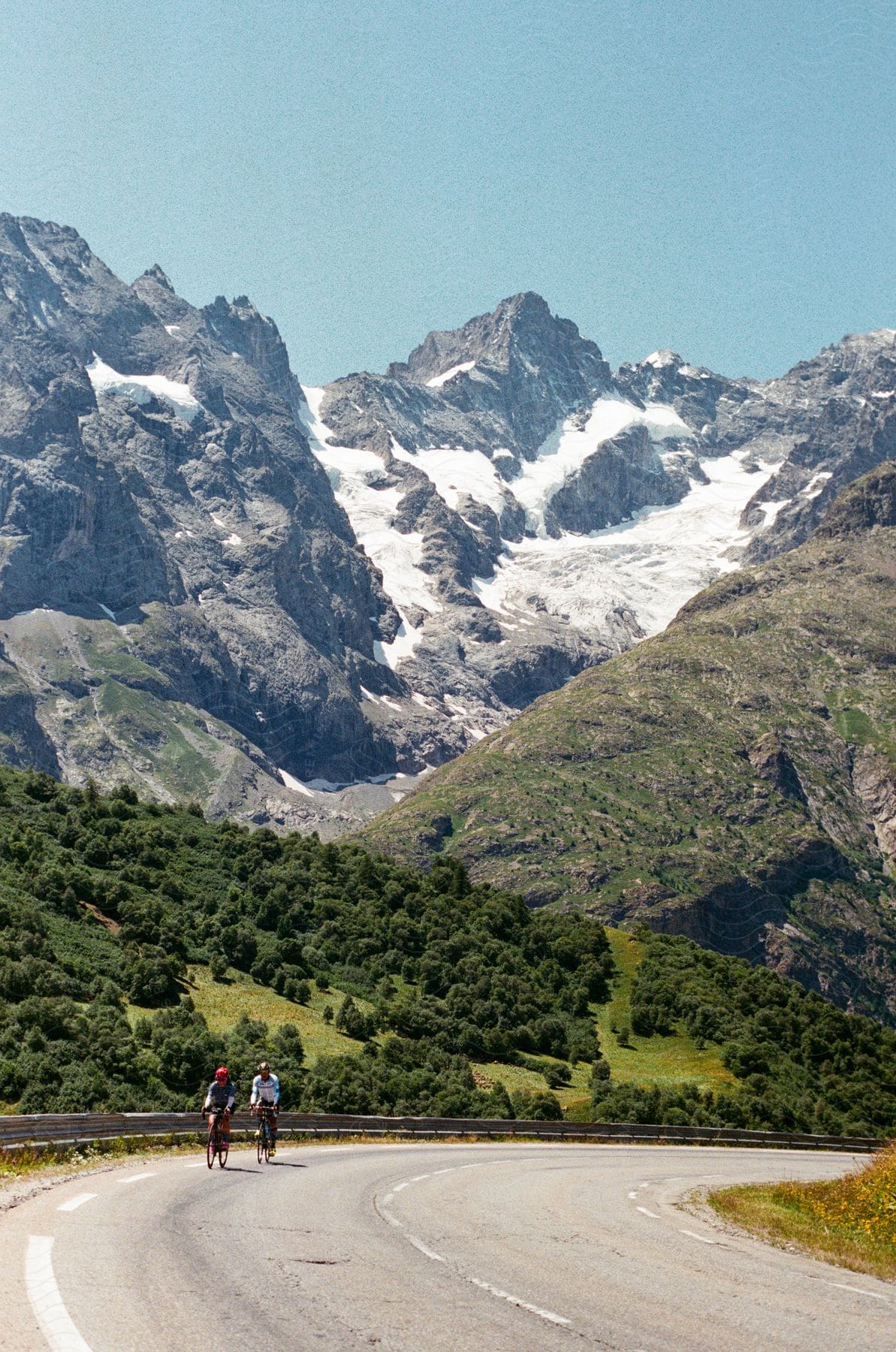 Two cyclists ride down highway at base of mountains.