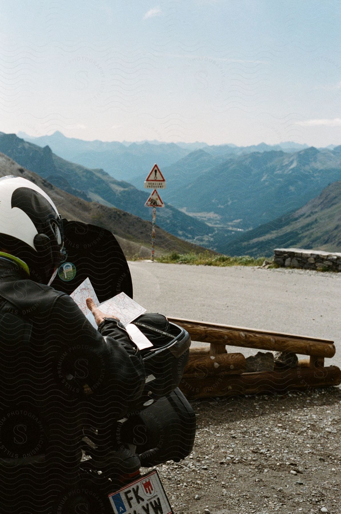 A lone motorcyclist studies their map, nestled on a mountain road enveloped by a backdrop of towering peaks and a roadside sign.