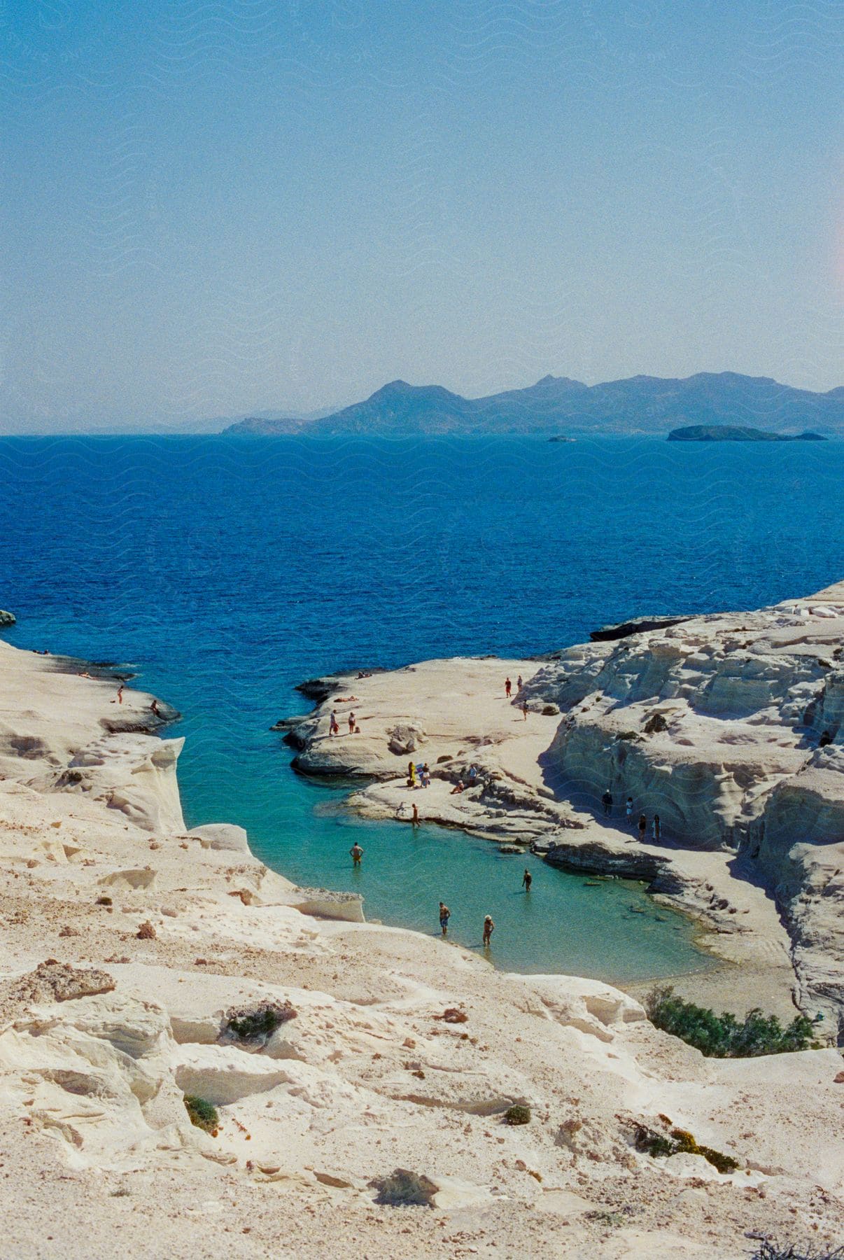 People are in the water and along the coast with mountains in the distance