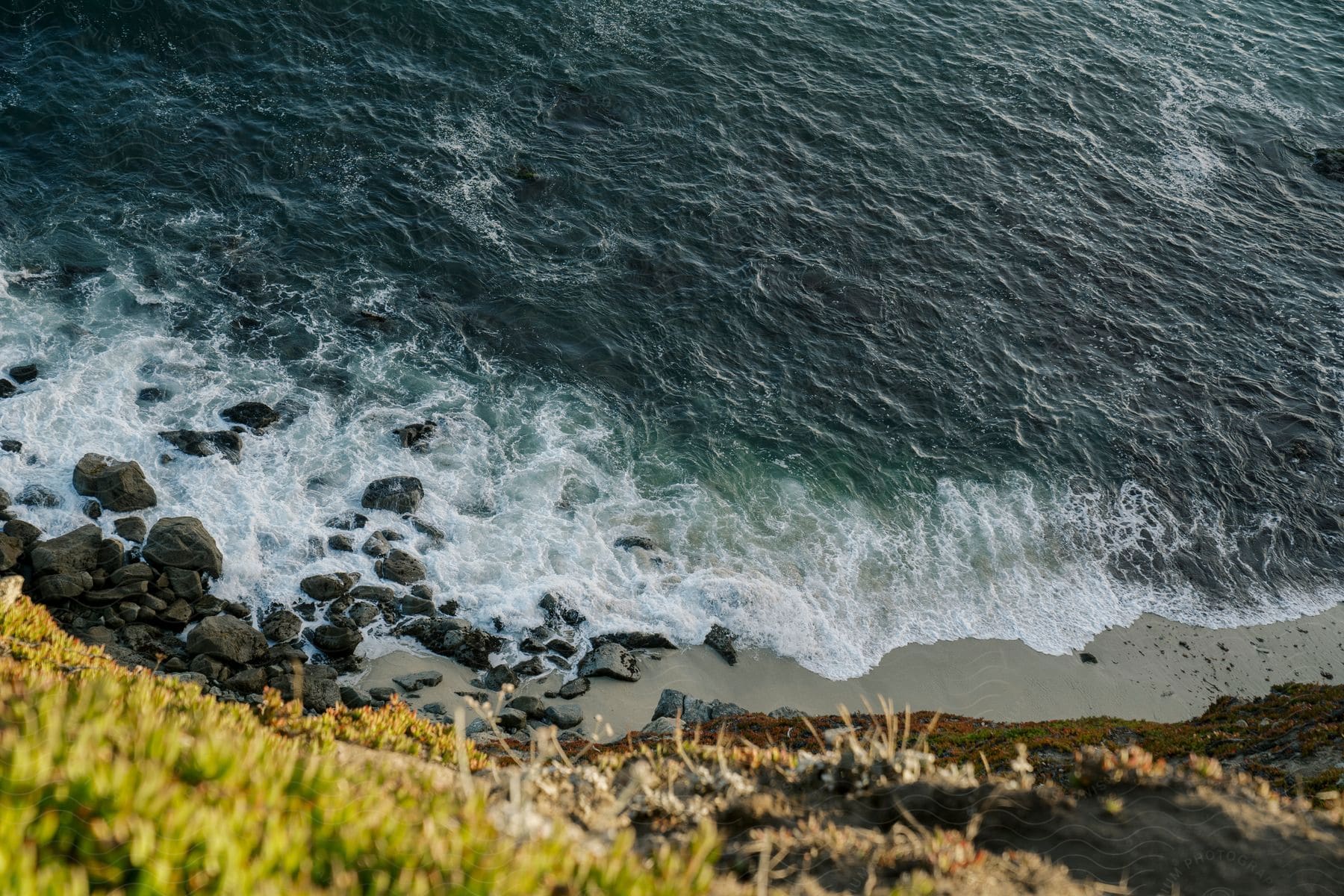 A grassy cliff overlooks a rocky shoreline as ocean waves wash upon the shore.